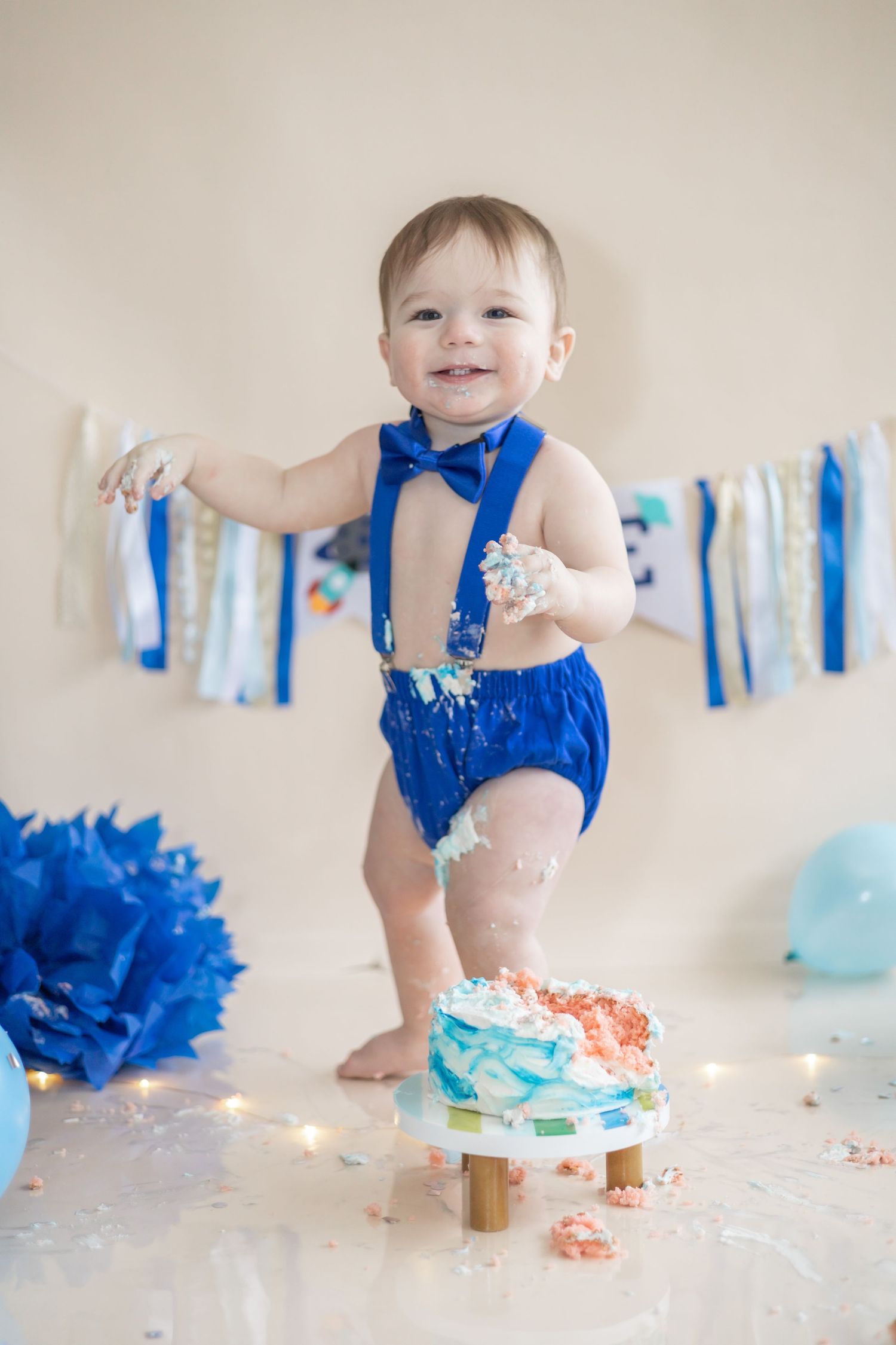 Baby in blue outfit and bowtie stands next to smashed birthday cake with blue decorations and string banners in background.