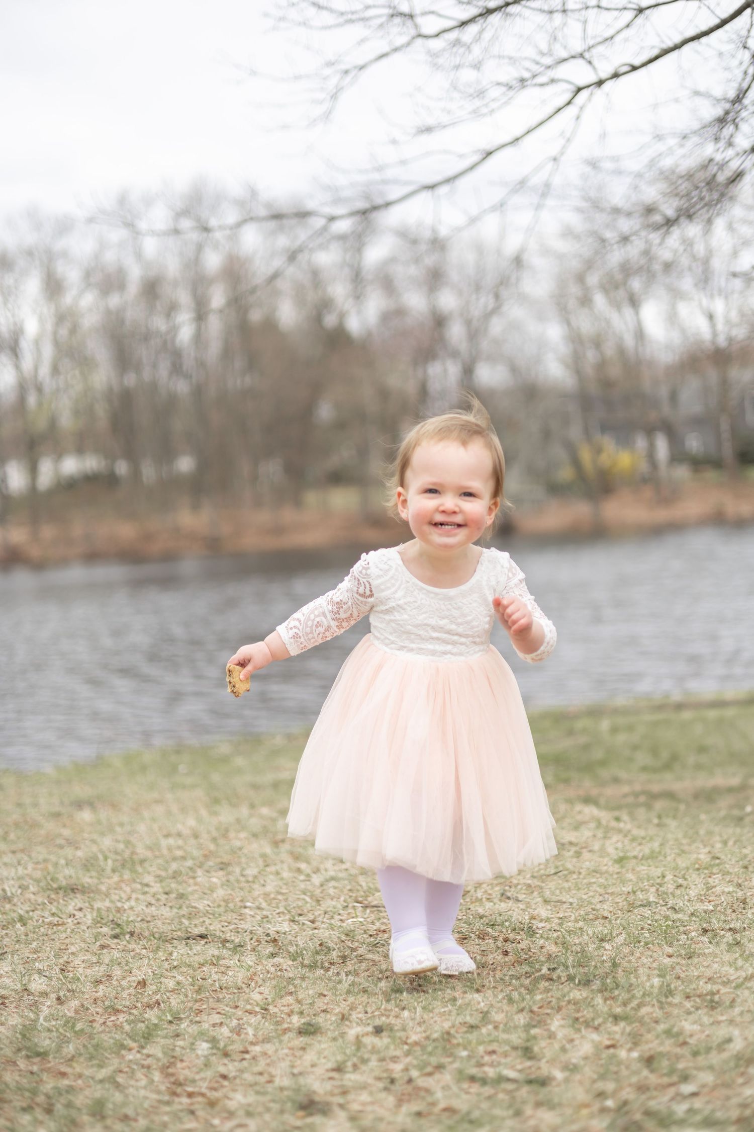 A small child in a pink tulle dress and white tights stands by a lake on a cloudy winter day.