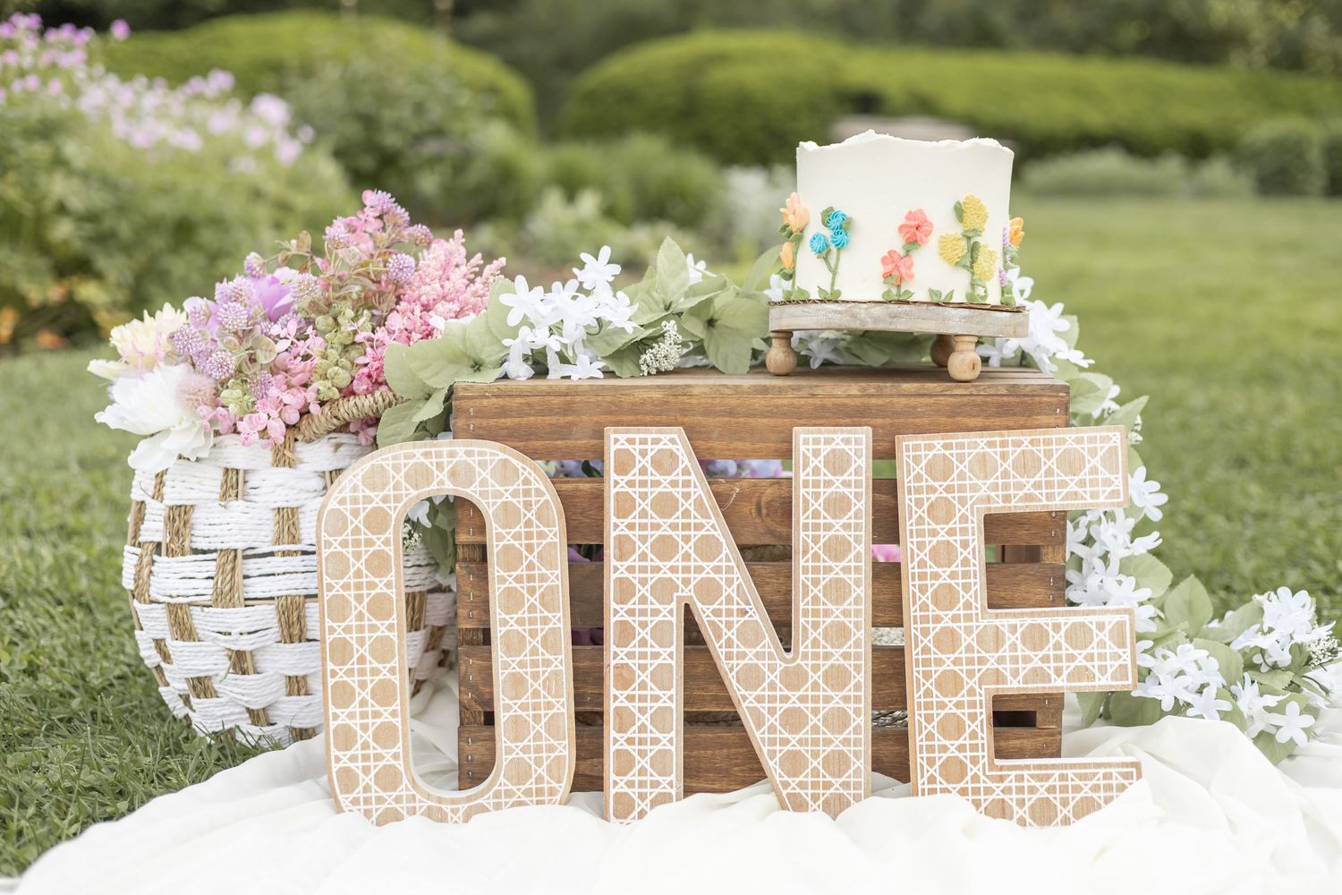 A decorative rustic first birthday display with marquee letters spelling ONE and a white birthday cake on wooden crates outdoors.