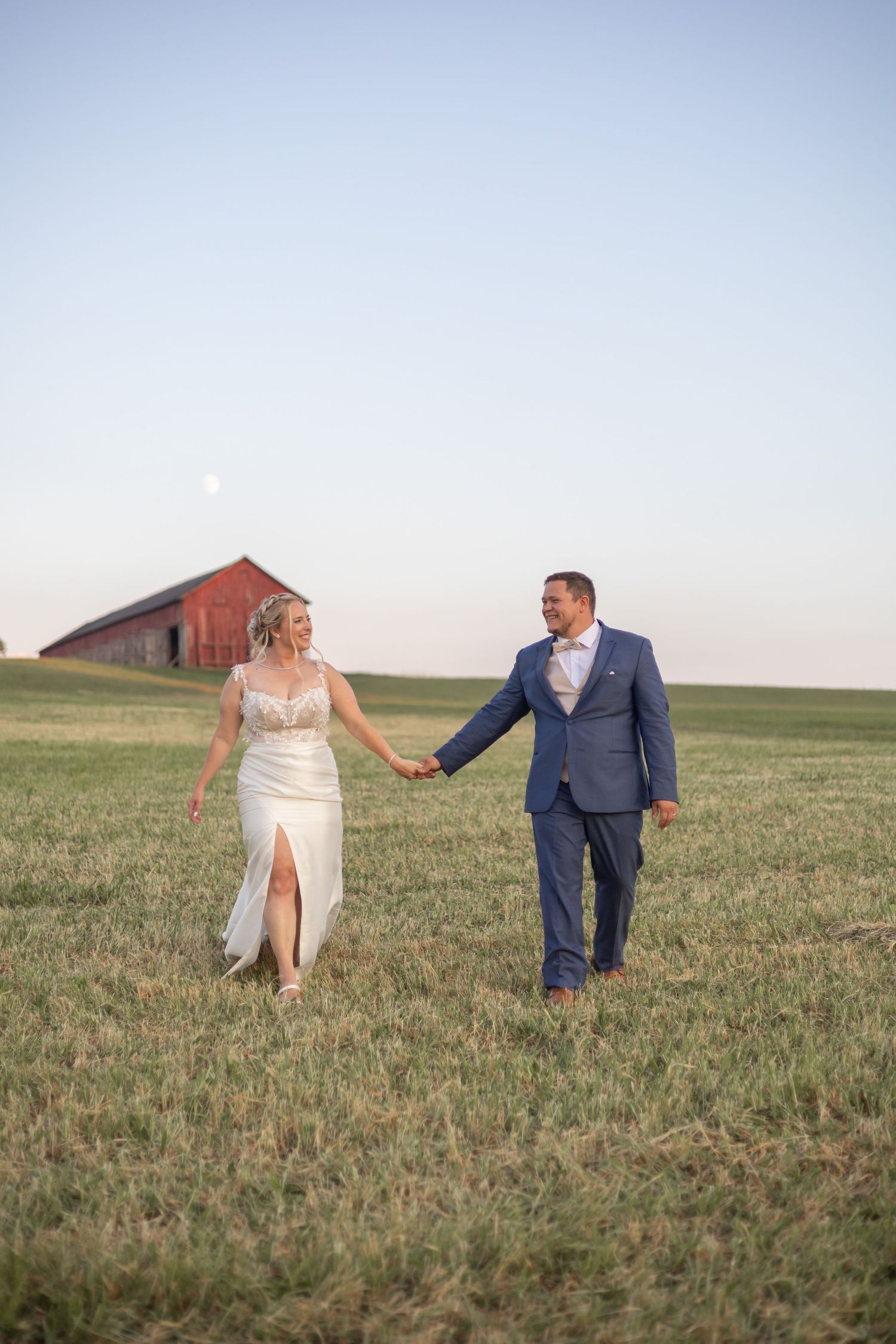 A couple walks hand in hand across a grassy field at sunset with a rustic red barn in the background.