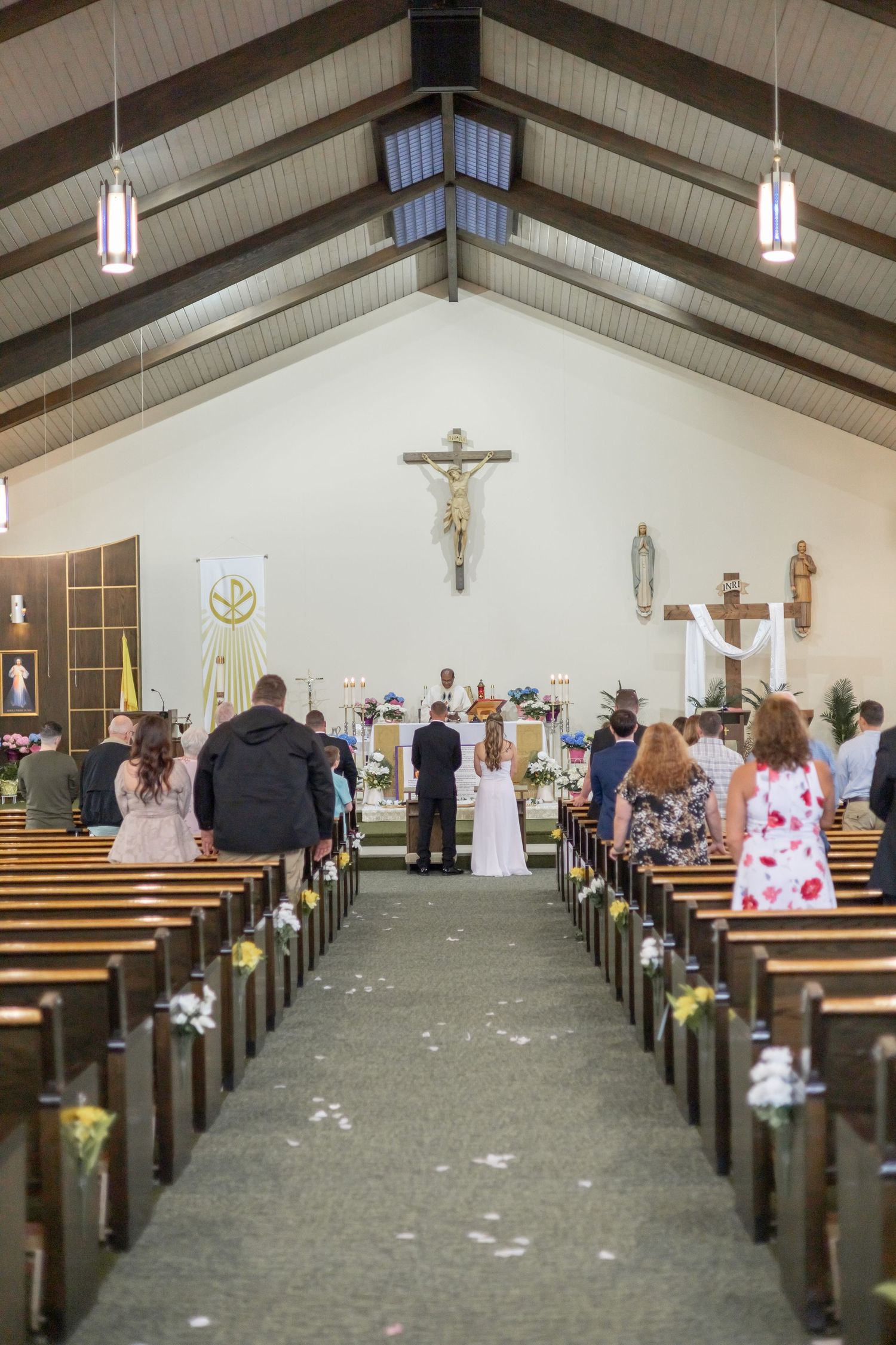 Interior view of a Catholic church wedding ceremony with wooden pews and scattered white petals along the aisle.