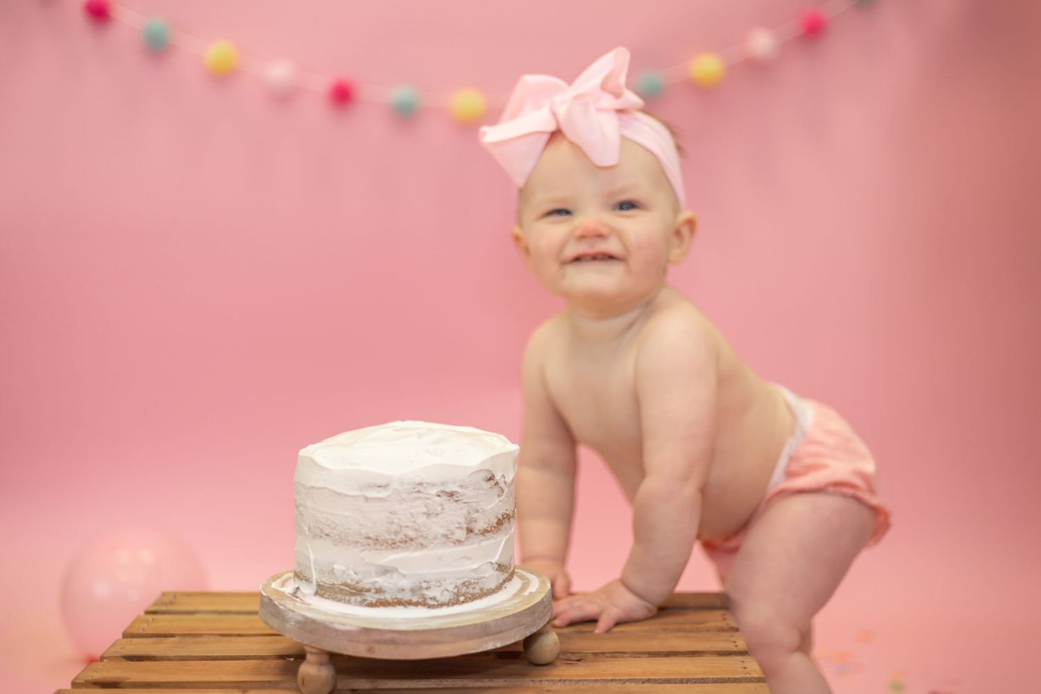 Baby in pink diaper and bow sits next to white cake during first birthday photo session against pink backdrop.