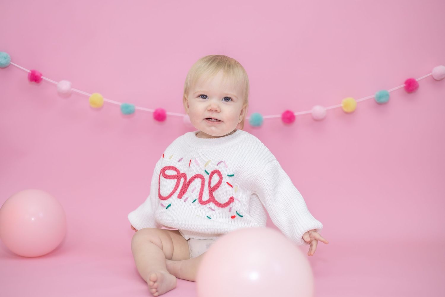 Baby in white one onesie poses against pink backdrop with colorful birthday party bunting decorations.