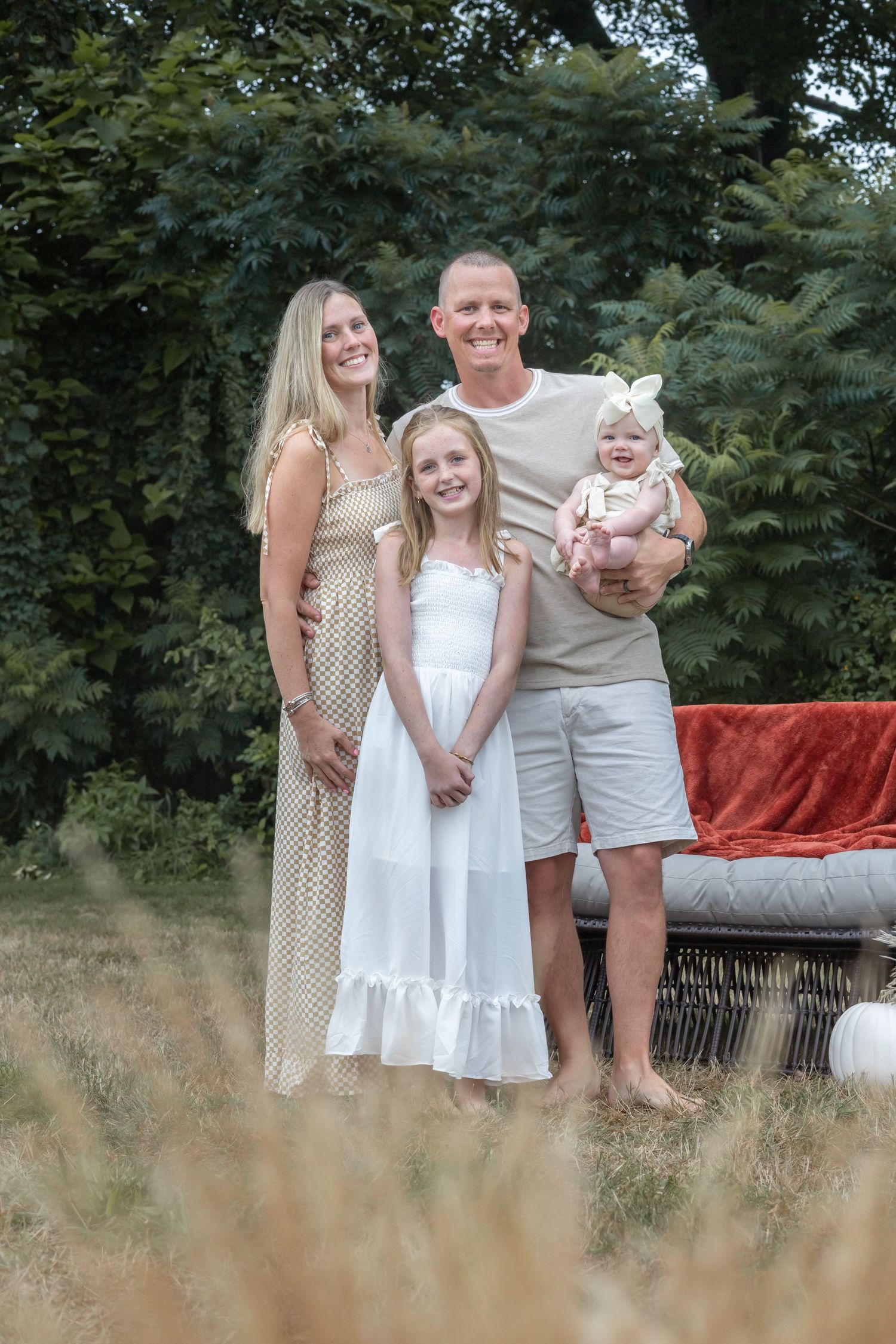 A family poses together outdoors in coordinated neutral outfits with a red cushioned seat behind them in a natural setting.