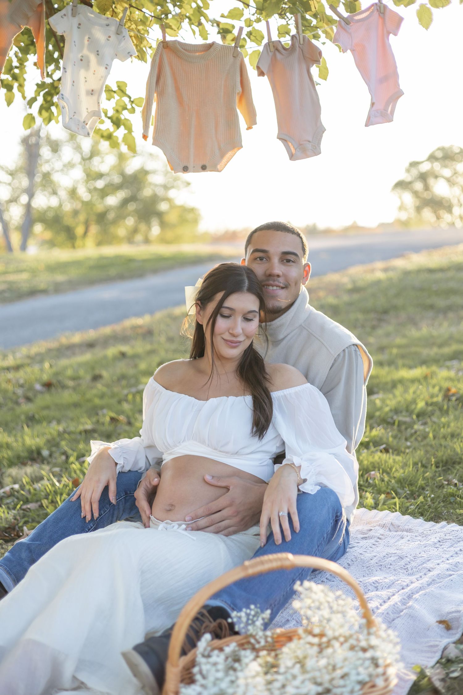 Maternity photoshoot in nature with baby clothes hanging on clothesline and couple sitting on blanket at sunset.