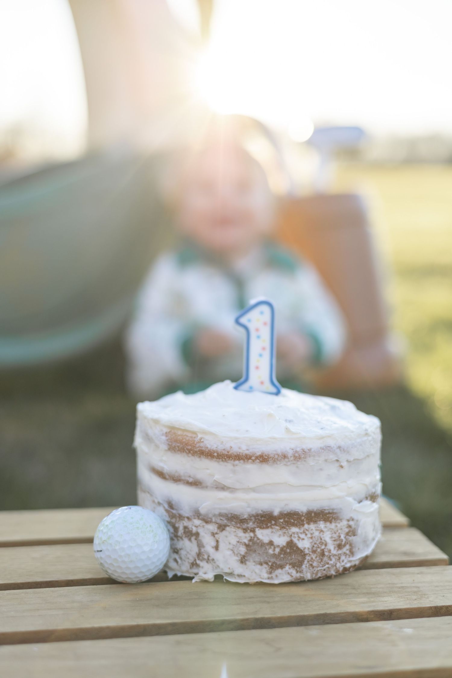 A small white frosted smash cake with a blue number one candle sits on a wooden table for a first birthday celebration.
