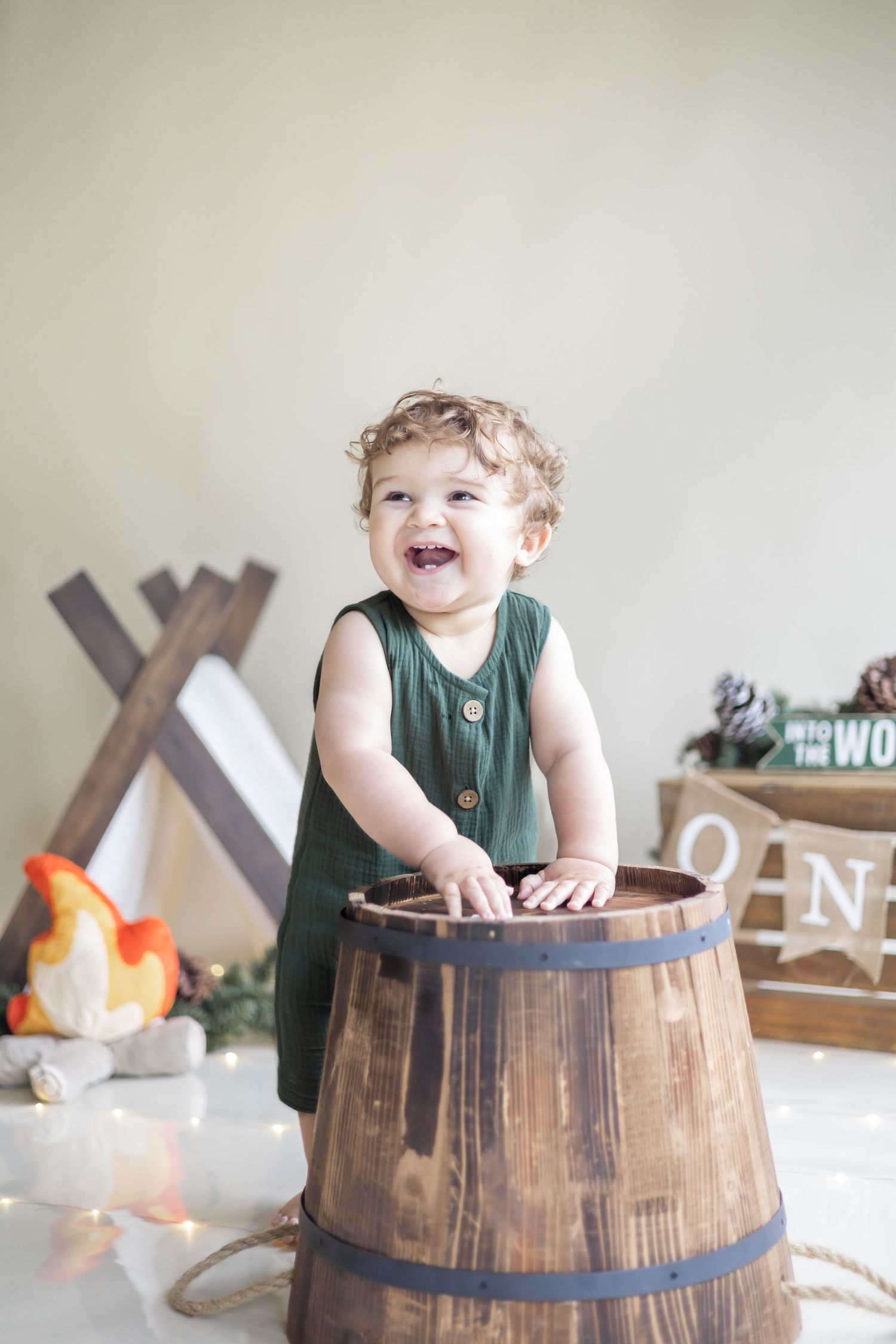 A joyful toddler in a green tank top leans on a rustic wooden barrel with decorative elements in the background.
