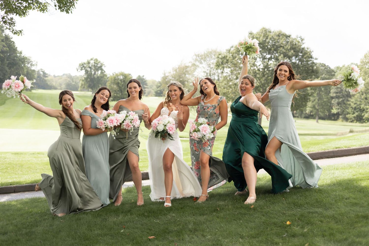 Bridesmaids in flowing dresses of sage green and emerald pose playfully on a grassy lawn during a wedding photoshoot.