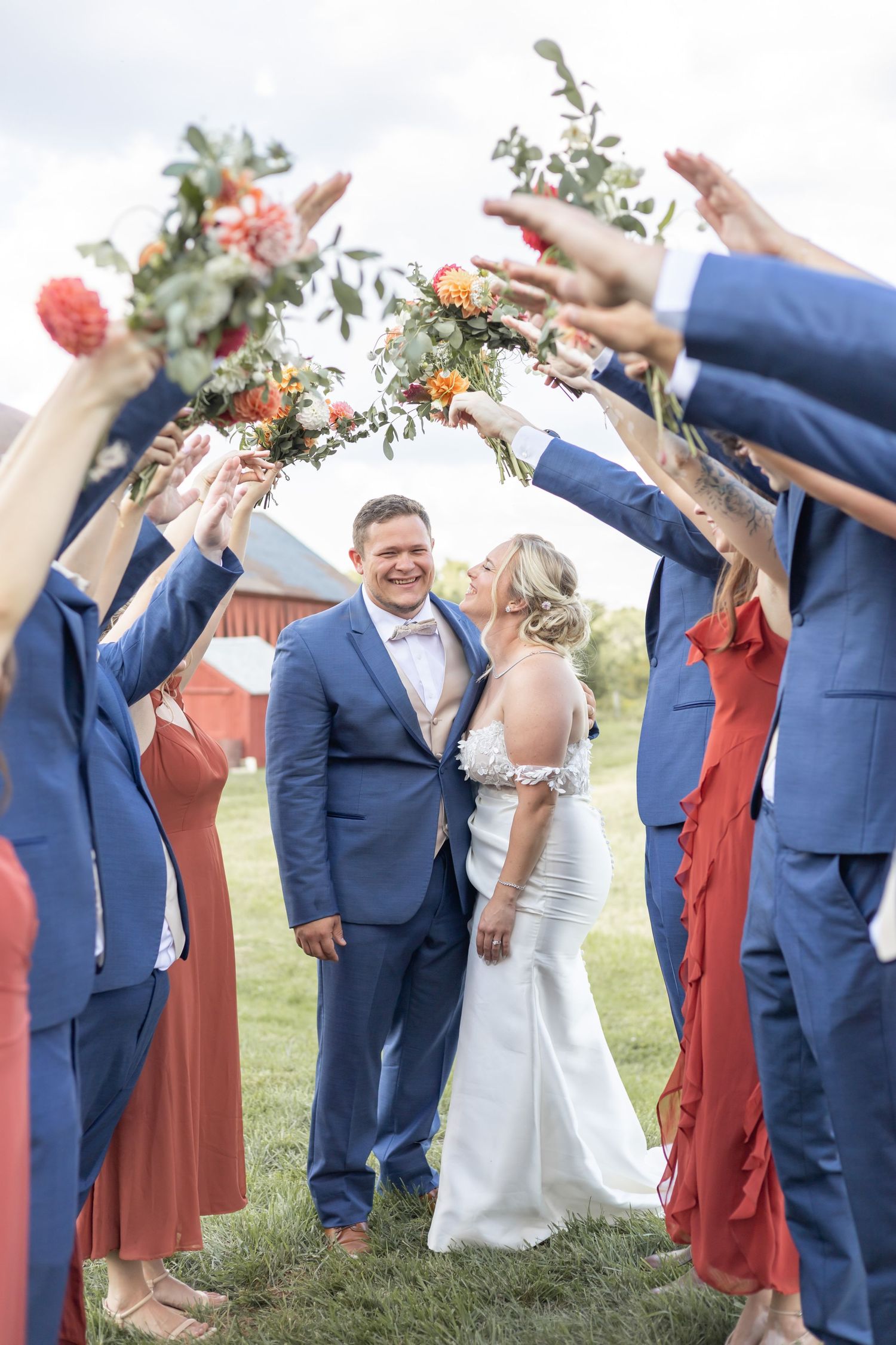 Wedding party in navy blue and coral attire creating an arch with floral bouquets on a grassy field by a red barn.