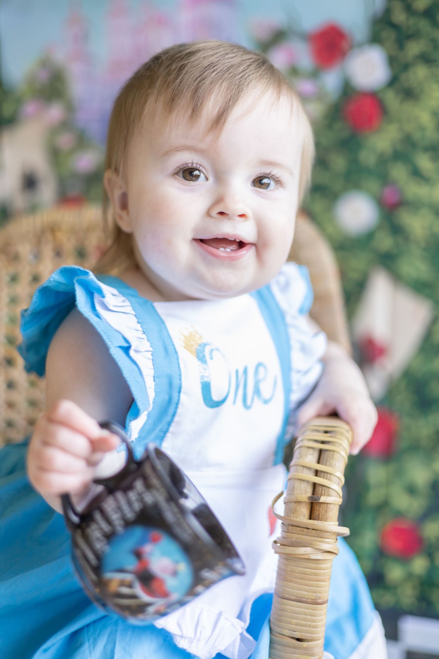 A smiling baby in a blue and white dress sits in a wicker chair against a floral backdrop.