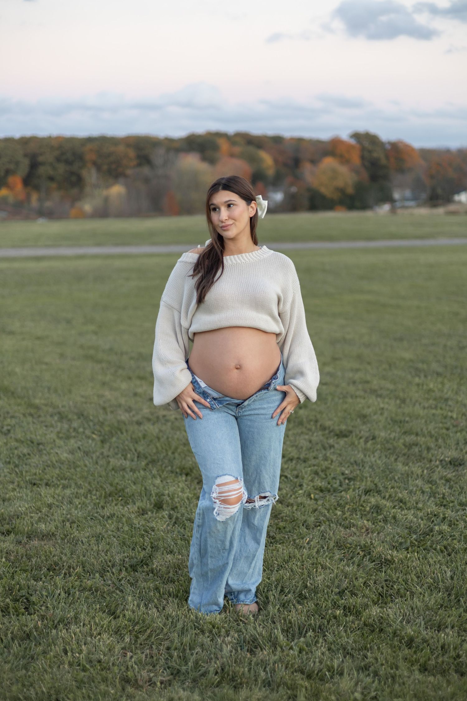 Expectant parent in cream sweater and distressed jeans poses in grassy field with autumn trees in background at sunset.