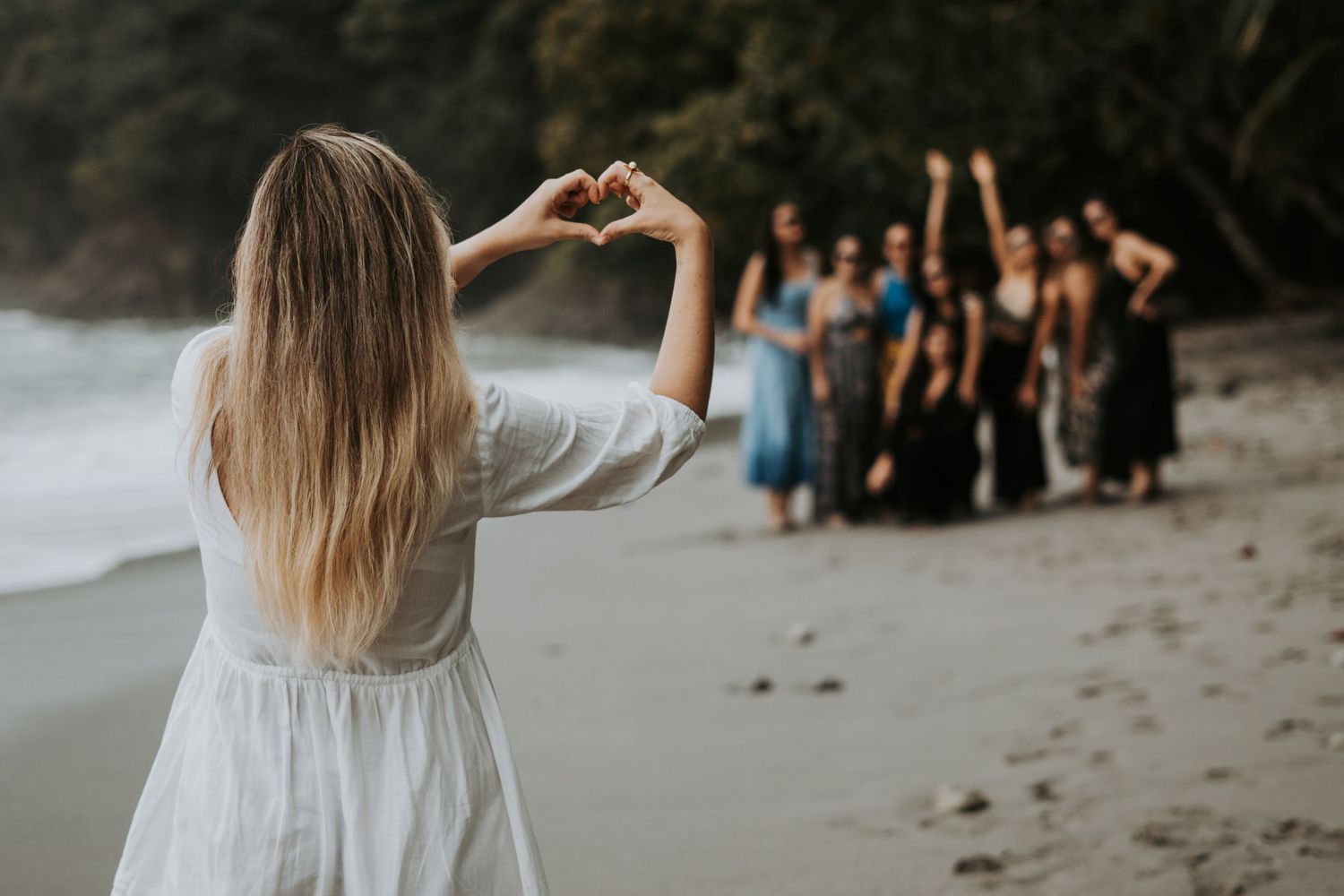 Woman in white dress making heart shape with hands toward group of friends on a tropical beach.
