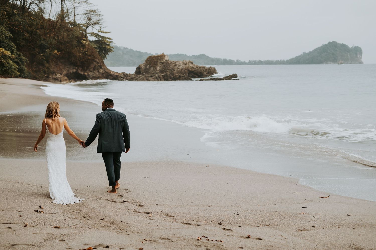 A couple walks hand in hand along a misty beach with rocky cliffs and ocean waves in the Pacific Northwest.