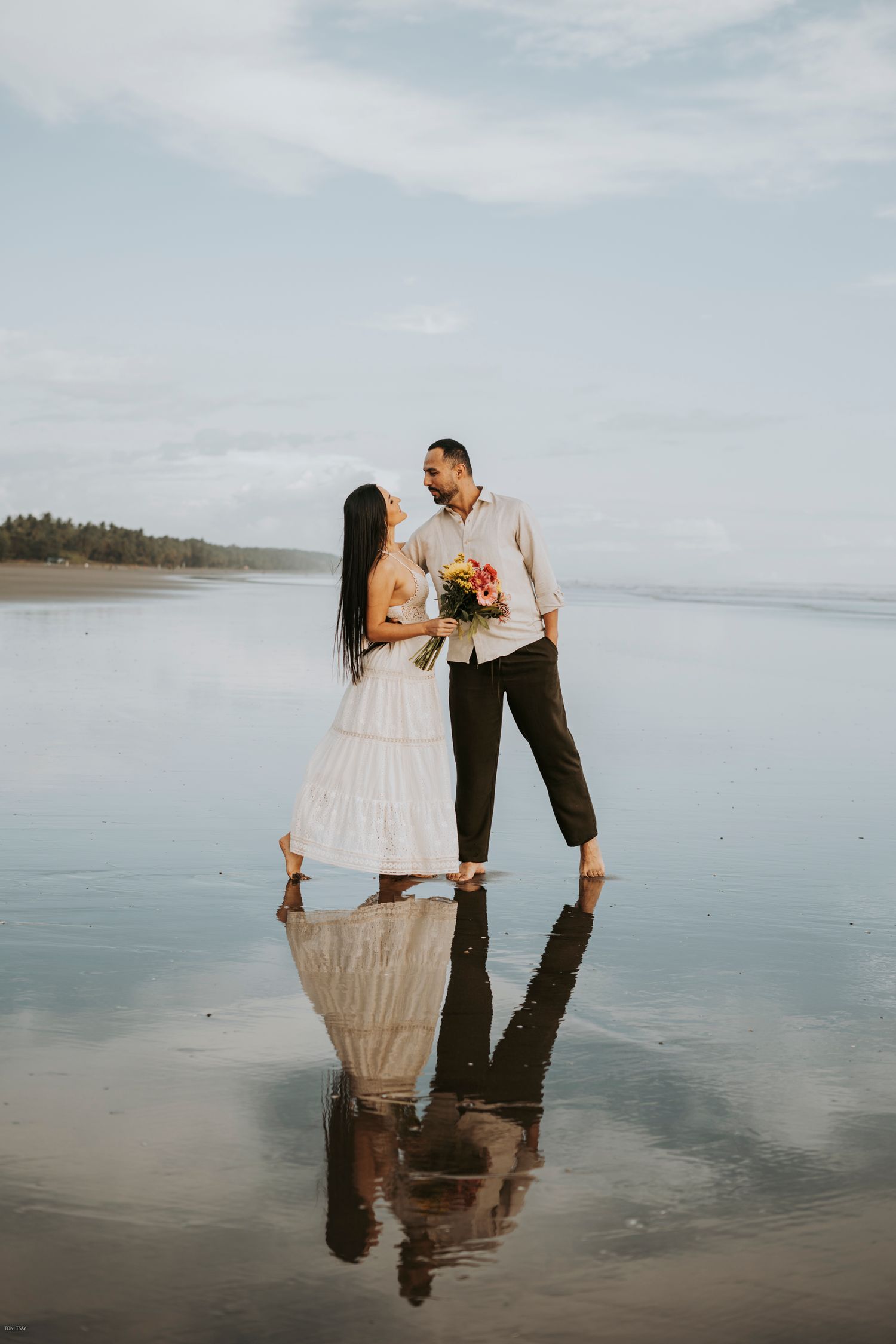 A romantic couple stands on a reflective beach at low tide with cloudy skies and distant shoreline in the background.