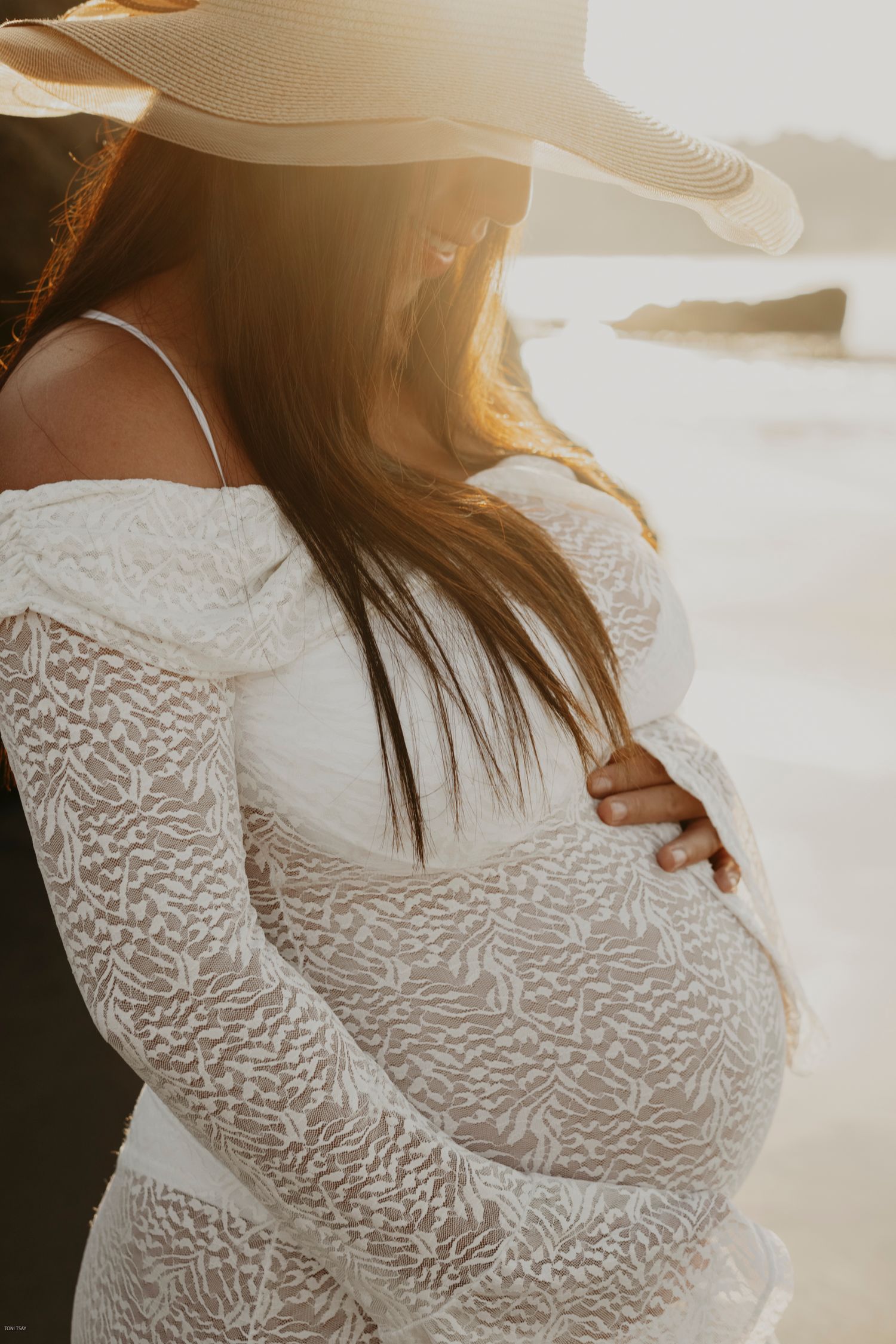 Pregnant woman in white lace dress stands in golden sunlight during maternity photoshoot at sunset.