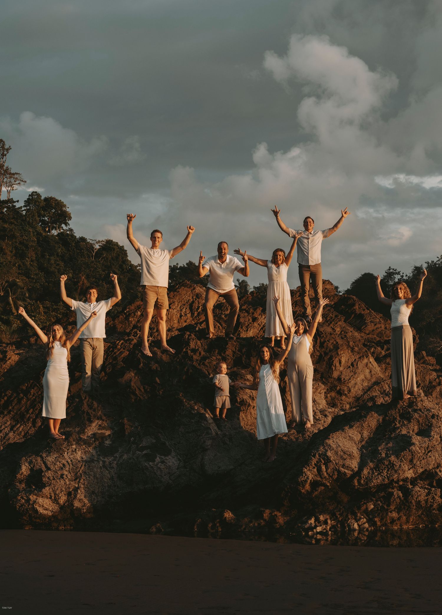 A group of people in white clothing celebrate on a rocky cliff at sunset, creating a joyful silhouette against the sky.