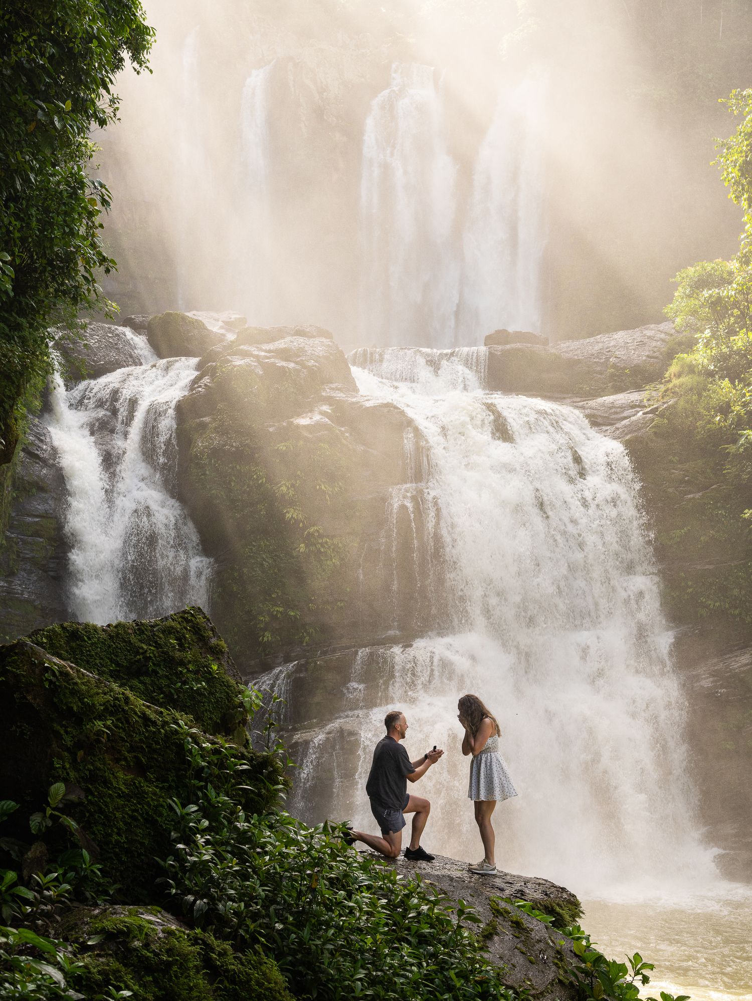 Silhouettes of people exploring a misty waterfall in a lush forest setting during golden hour.