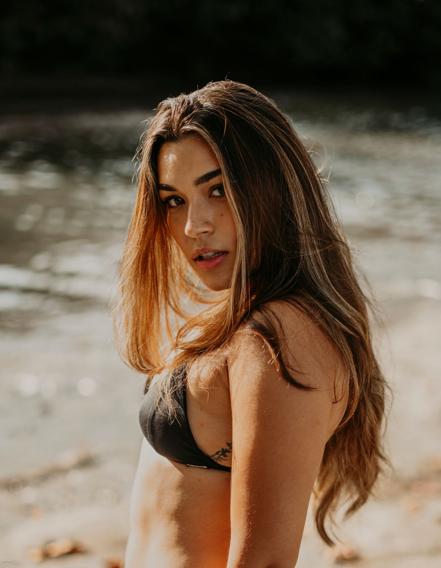 Person with long wavy brown hair poses in a black swimsuit on a beach with soft lighting and blurred background.