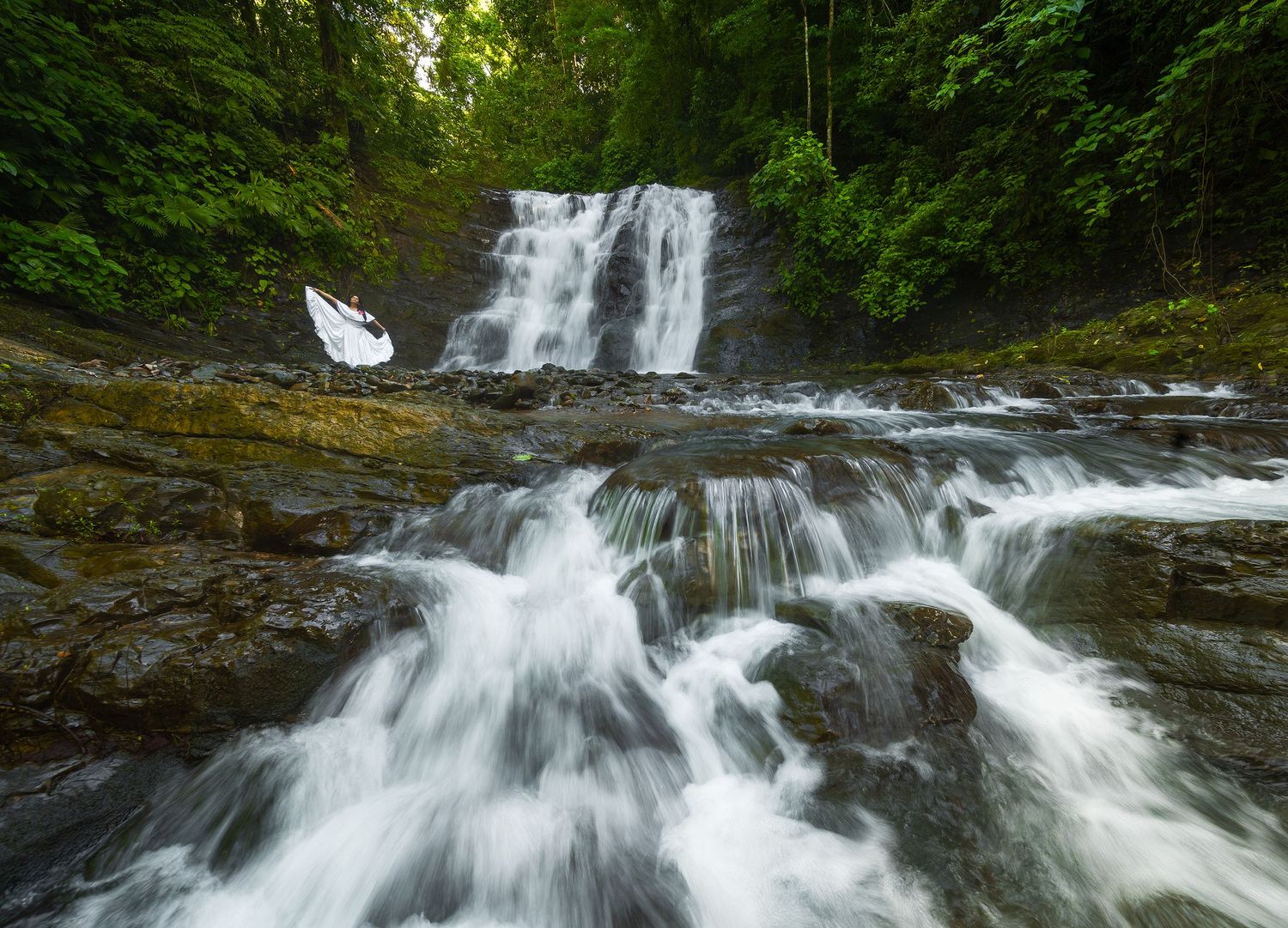Multi-tiered waterfall cascades through lush green forest with smooth flowing water.