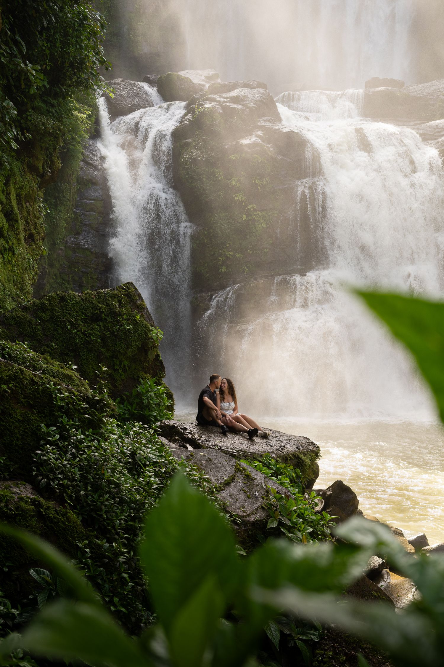 Couple sits on rocky outcrop in Nauyaca waterfall surrounded by lush greenery in misty tropical setting.