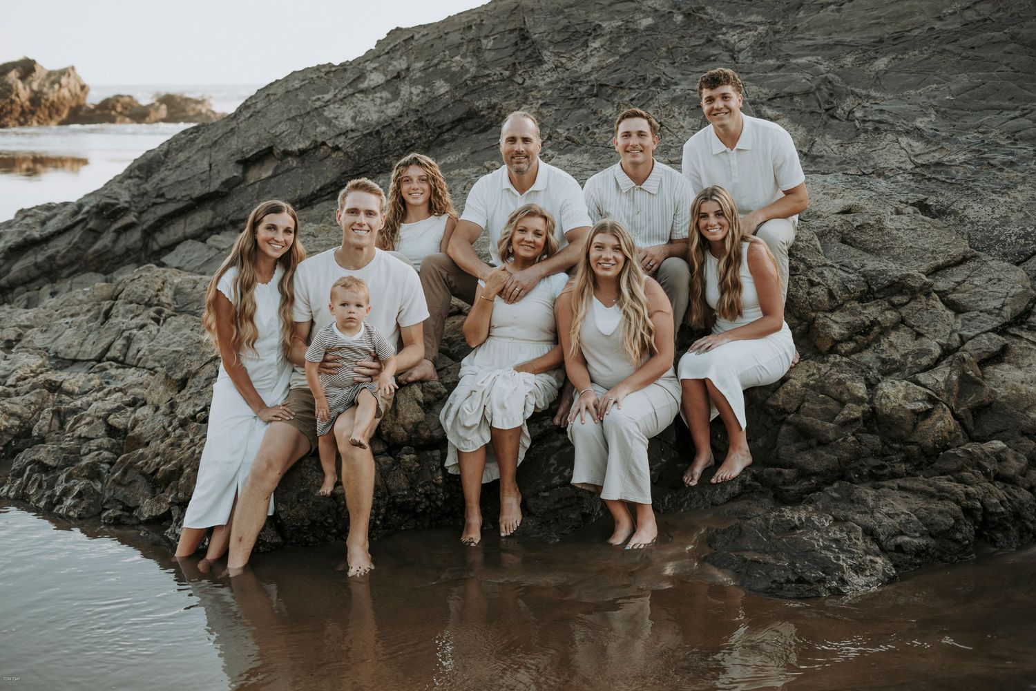 A group in white outfits sits together on dark coastal rocks by the ocean at sunset.
