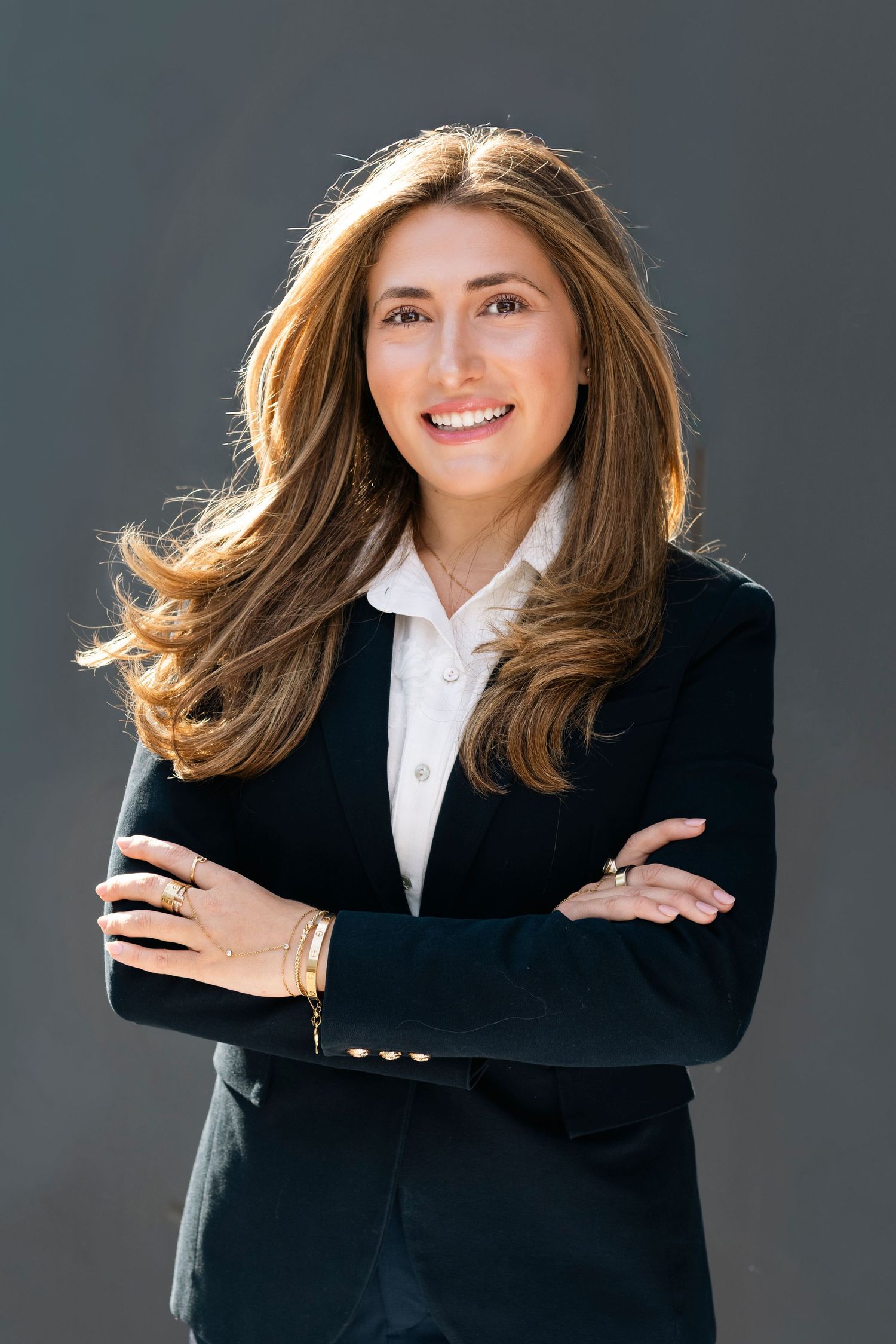 Professional businesswoman in black blazer smiling with arms crossed against gray background.