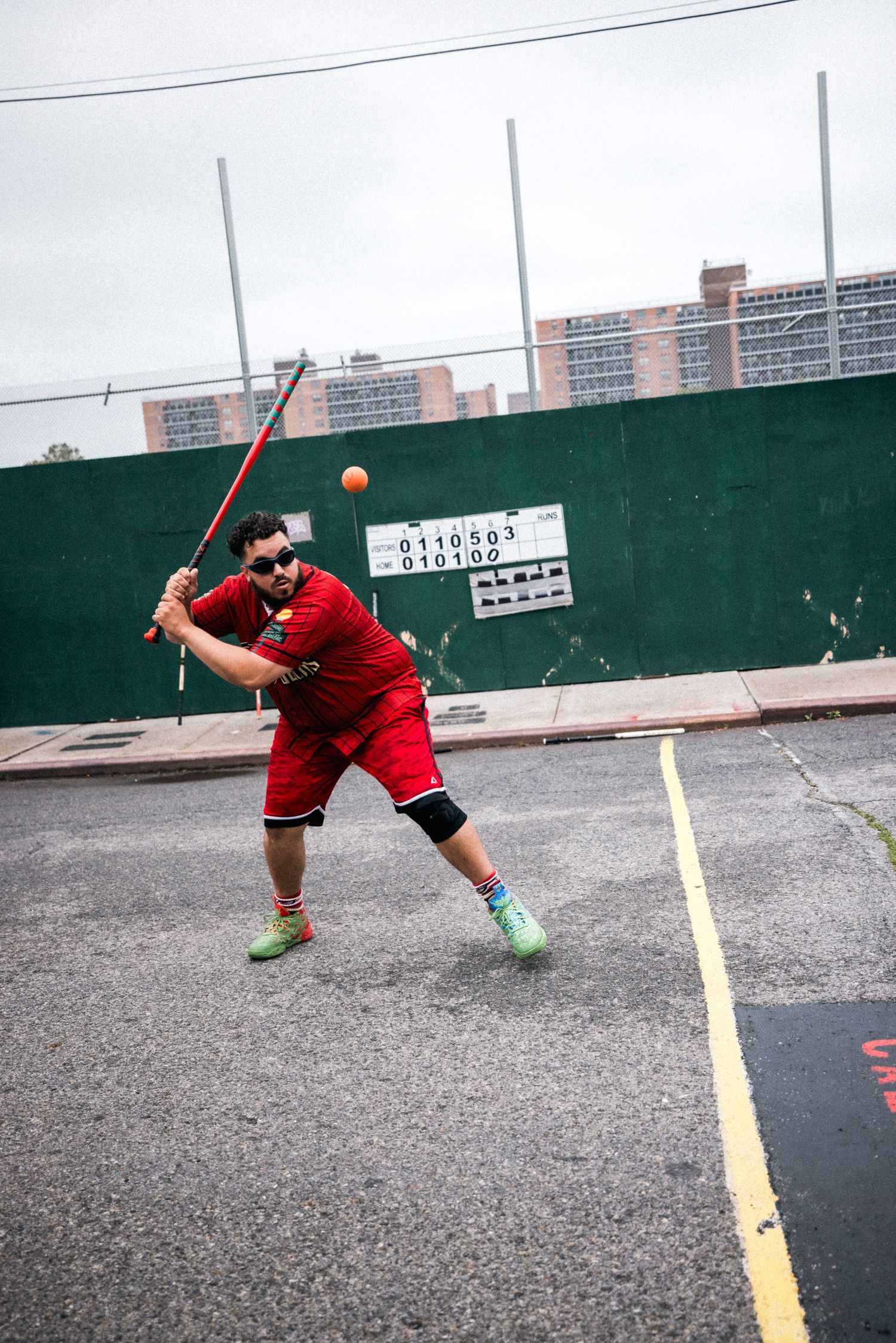 Player in red shorts demonstrates dynamic stickball batting stance sequence on urban court with green wall.