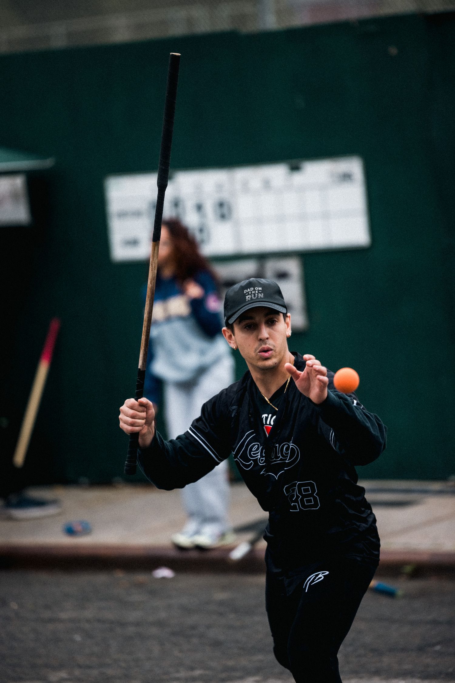 Athletic player in dark uniform swings a bat at a ball in an urban setting.