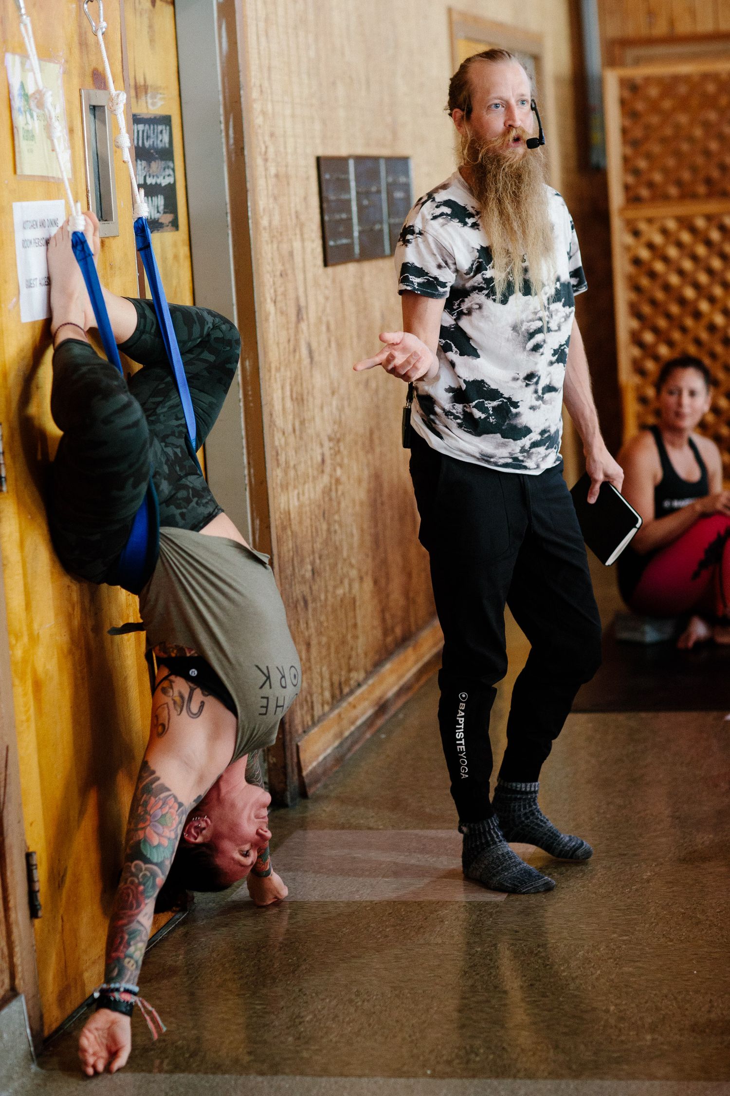 Person practicing aerial yoga while an instructor in tie-dye shirt gestures nearby.