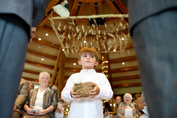 Young ring bearer in white holds a pillow under hanging decorations during a wedding ceremony.