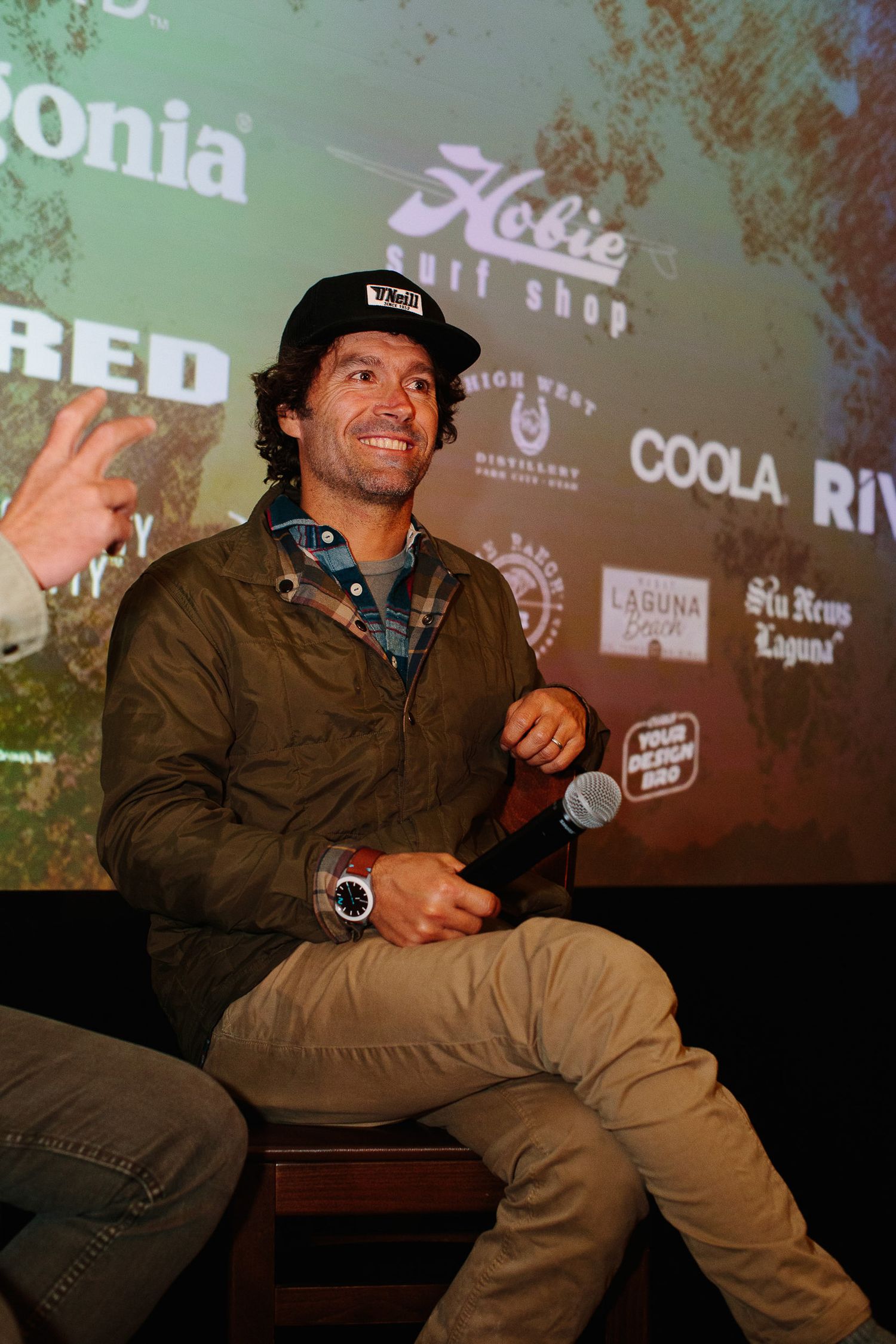 Speaker on stage at an event with projection screen showing outdoor brand logos in background.