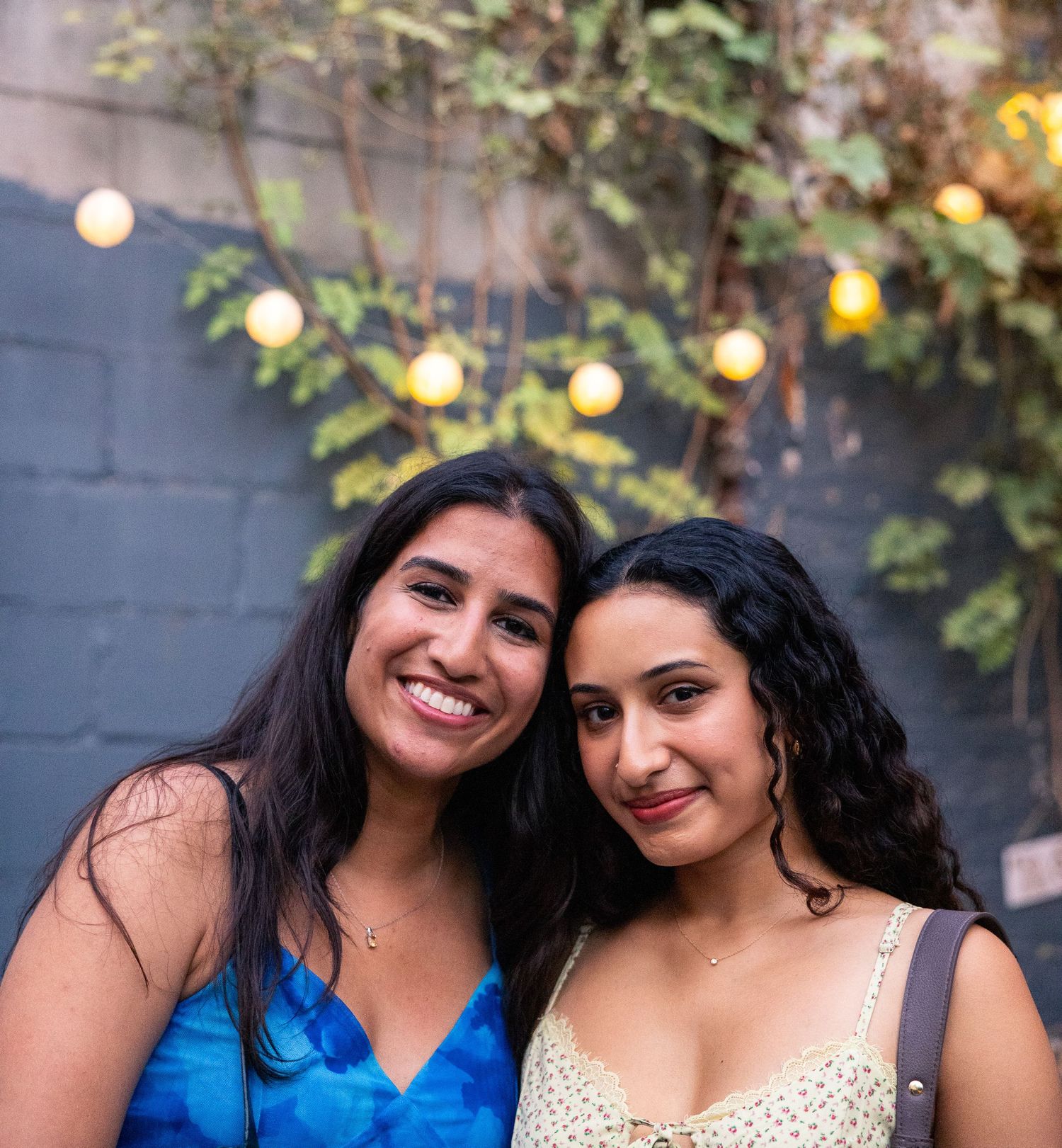Two women smile at an outdoor party with string lights and foliage in the background.