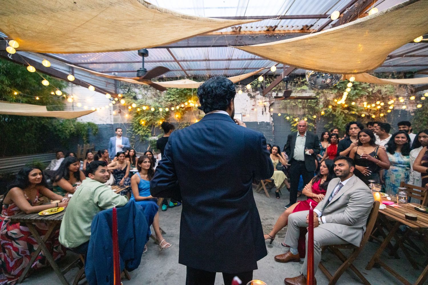 A speaker addresses seated guests at an outdoor event under string lights and shade sails.
