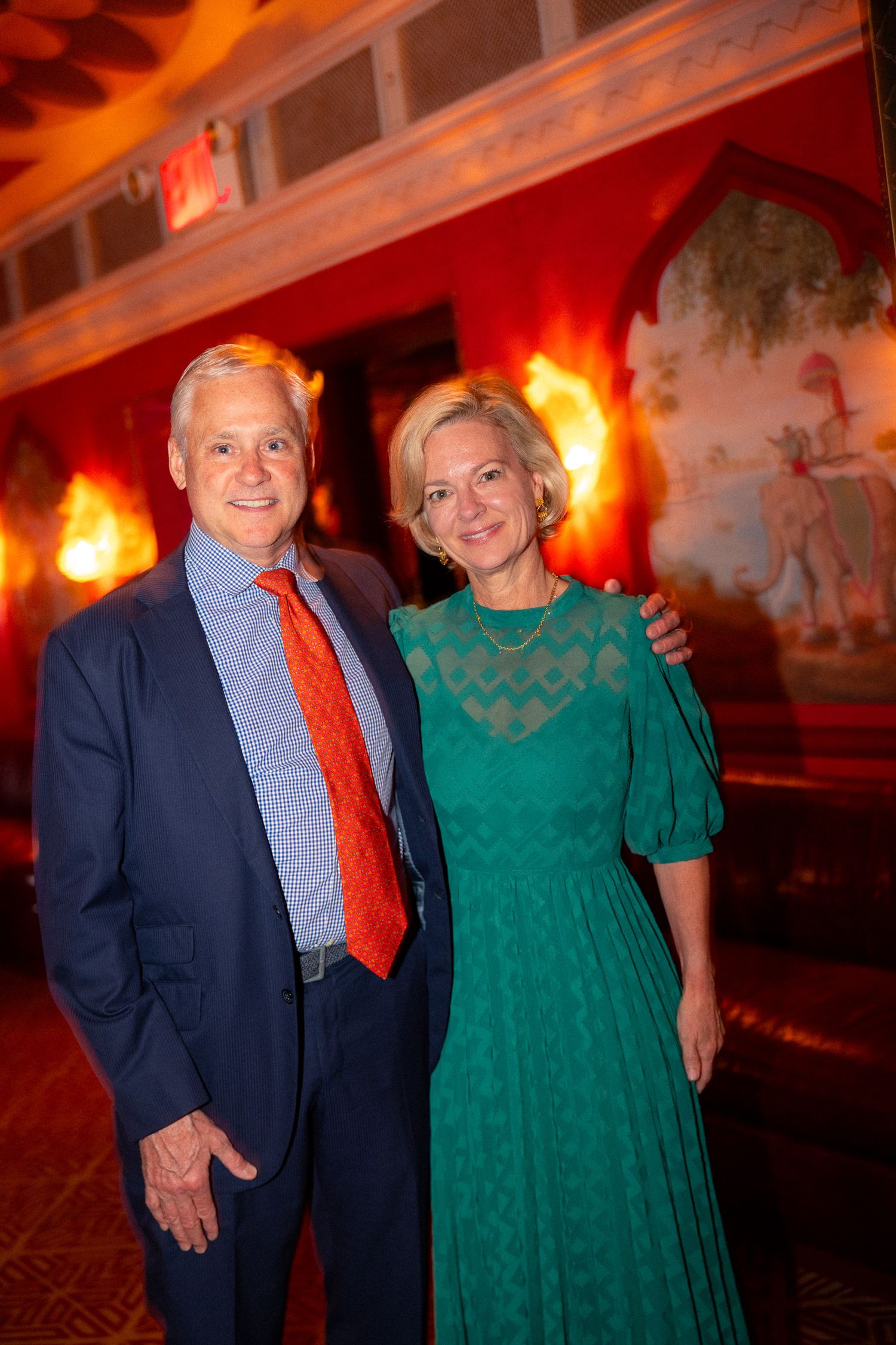 Couple in formal wear posing against a red-lit wall with decorative murals.
