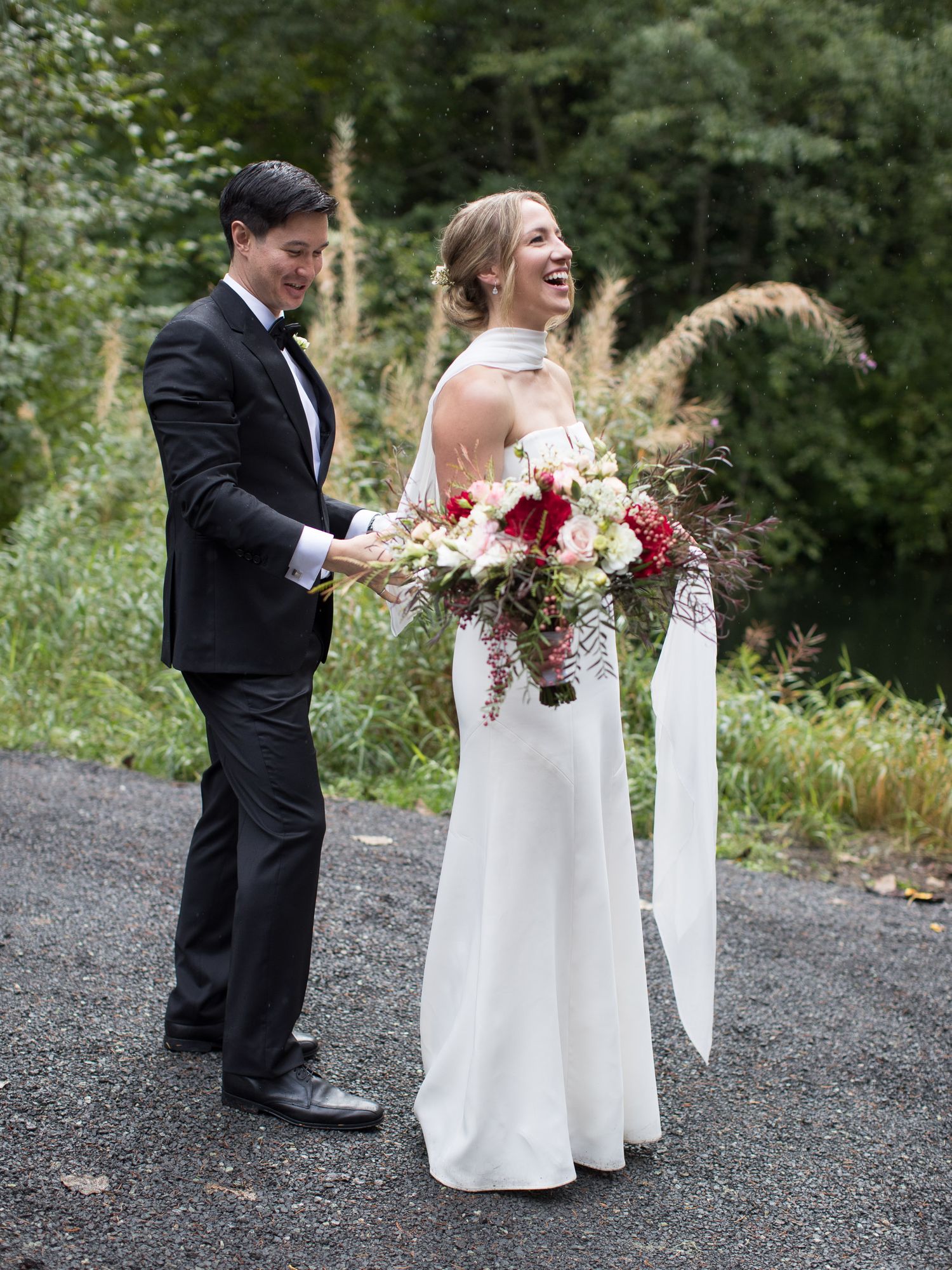 A couple in formal wedding attire stands on a gravel path during their outdoor photoshoot.