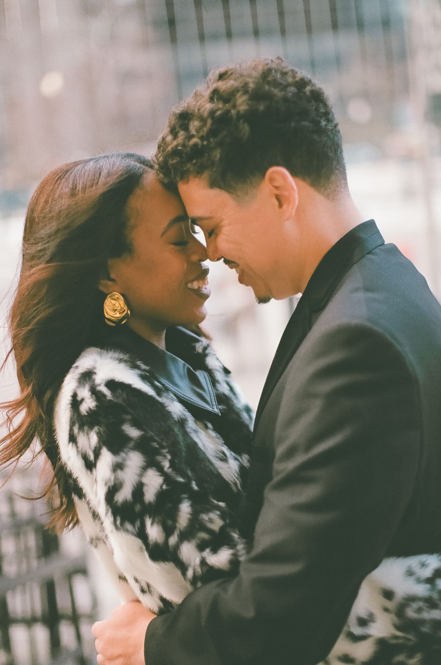A couple shares a romantic moment, touching foreheads and smiling, dressed in elegant attire outdoors.