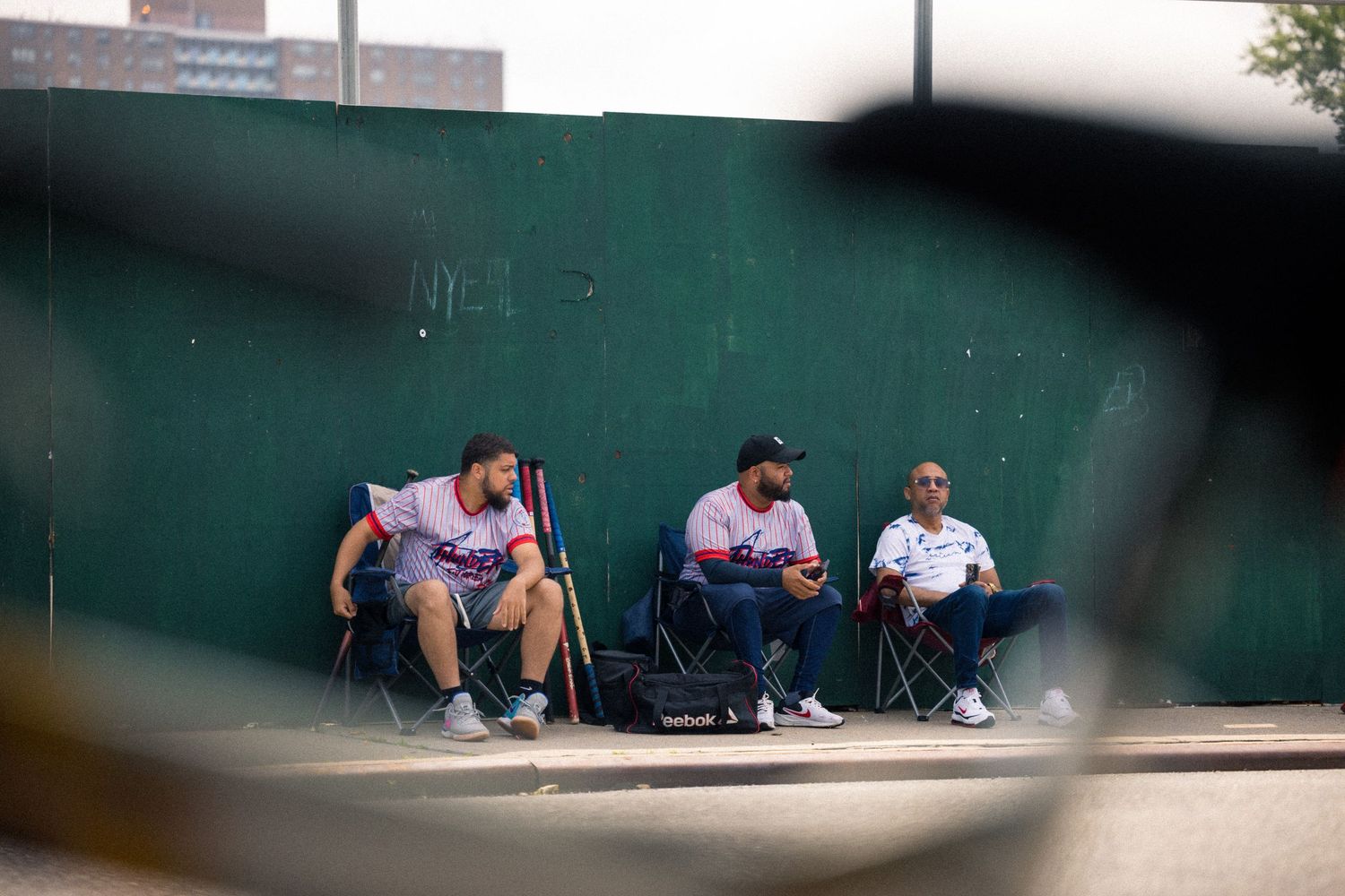Three people sit in camping chairs along a green wall while waiting outside in casual clothes.