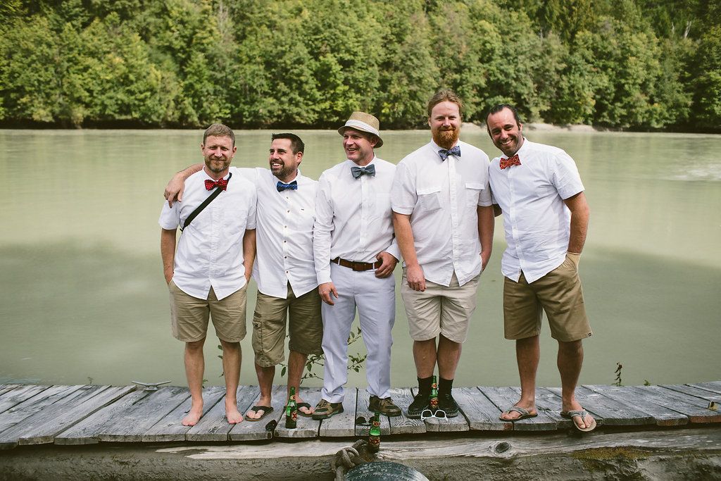Groomsmen sitting together on a dock by a lake wearing khaki pants and white shirts.