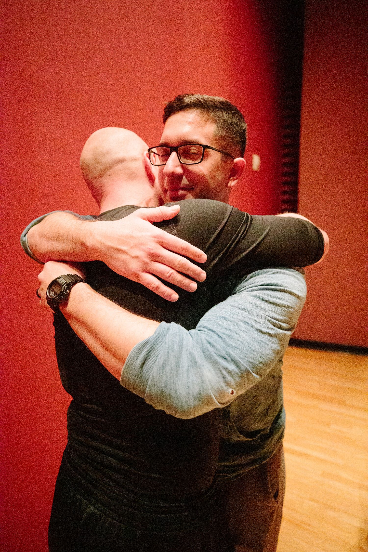 Two people sharing an emotional embrace in a performance space with red walls.
