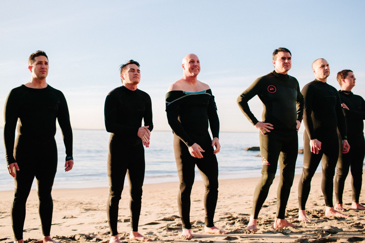 A line of people in black wetsuits standing on a sunny beach.