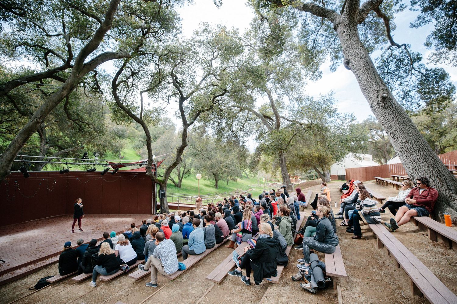 A large group of students sit on wooden amphitheater-style benches under shady trees during an outdoor gathering.