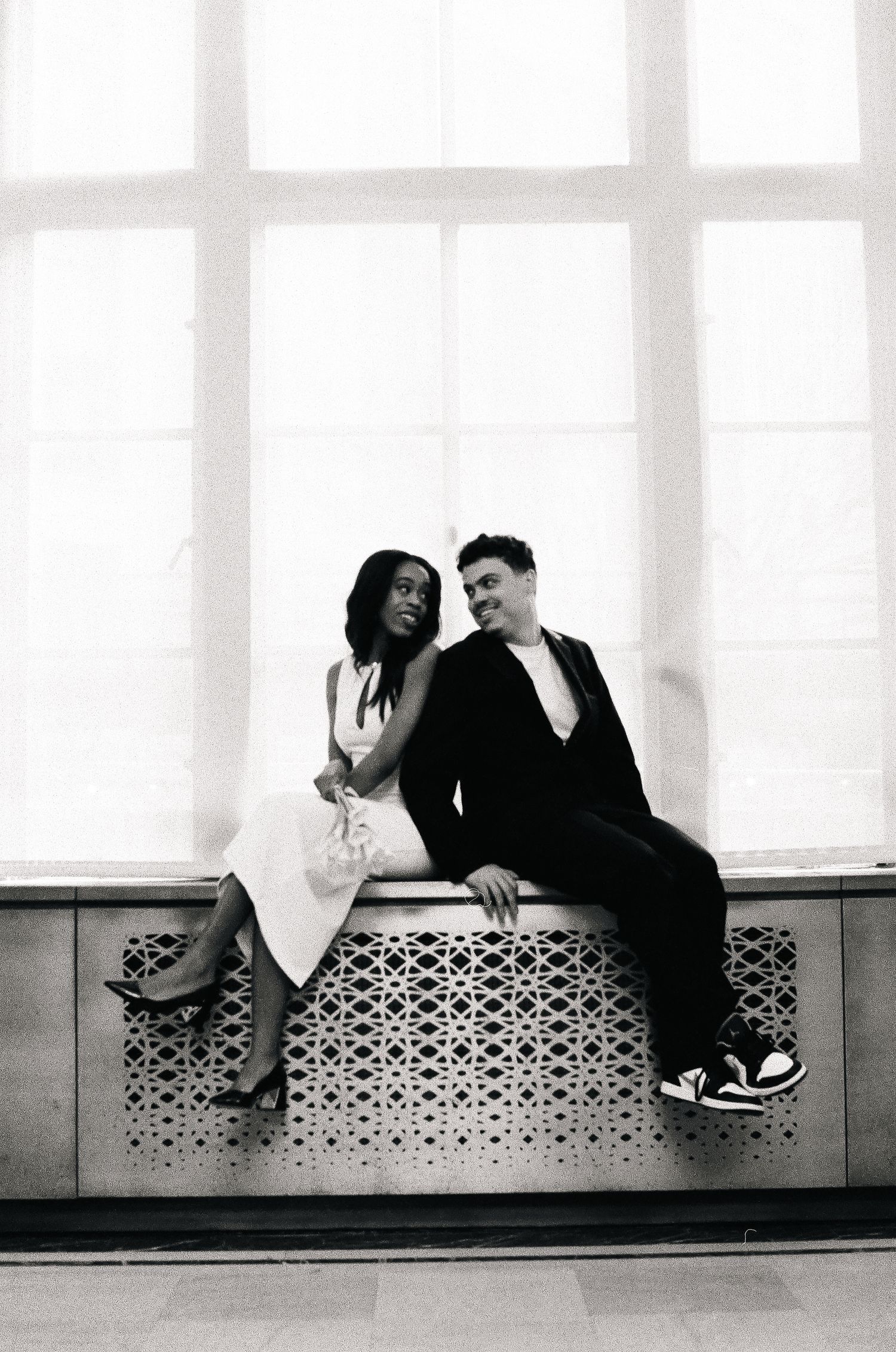 Black and white photo of a couple sitting together on a decorative ledge by a large window.