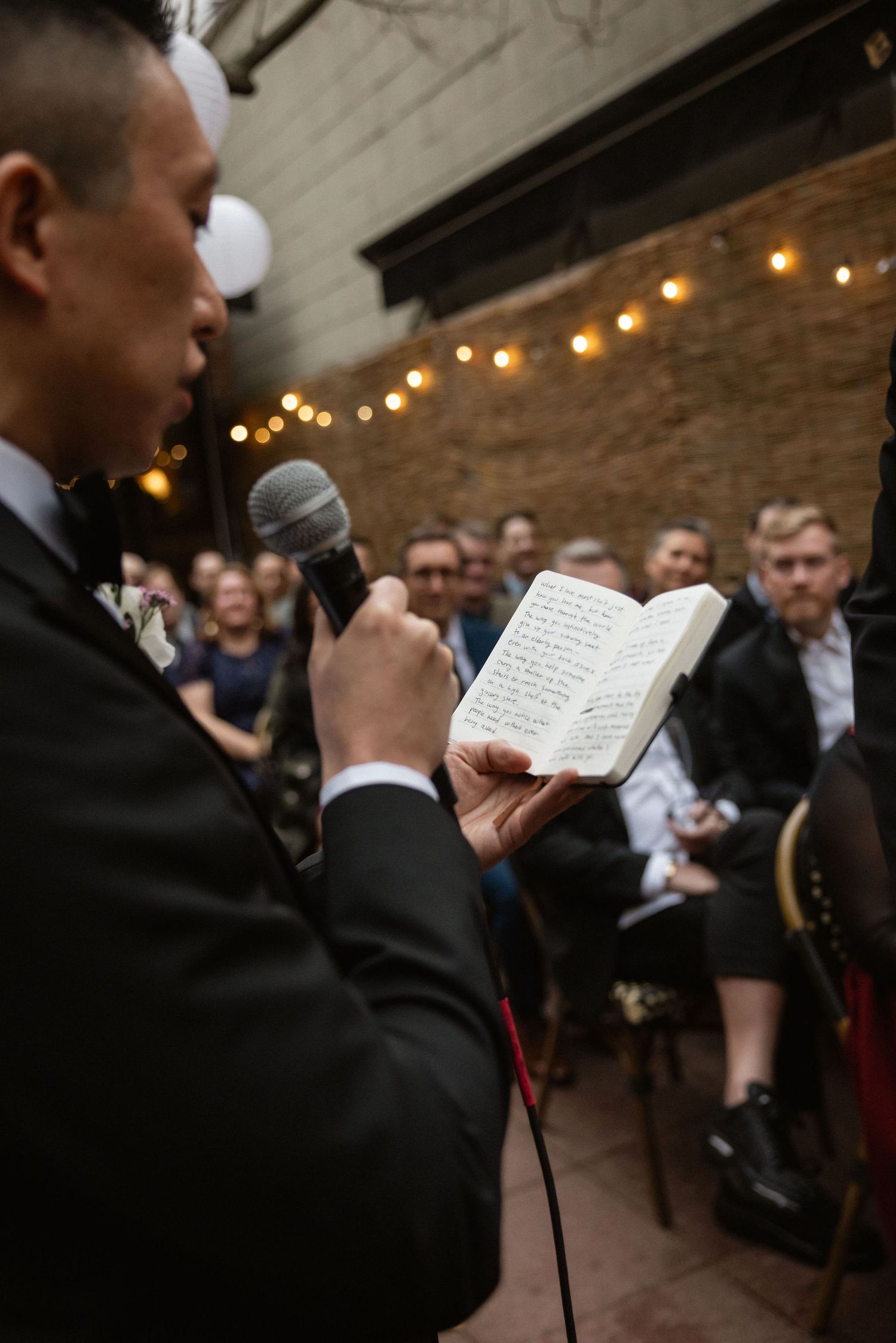 Groom reads vows from a notebook while holding a microphone at an indoor wedding ceremony with guests seated behind.