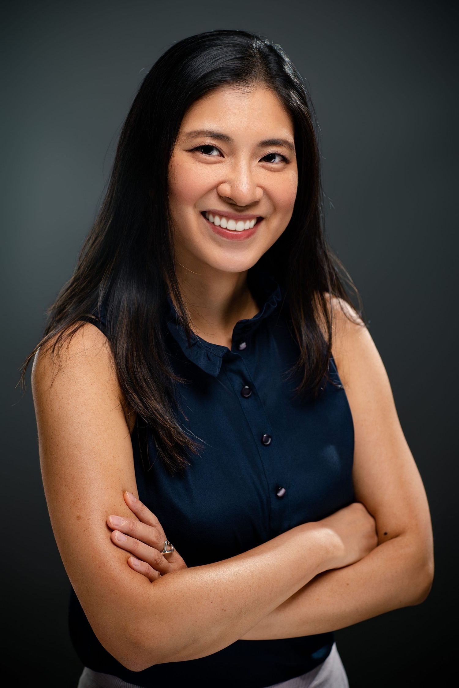 Smiling Asian woman in navy sleeveless blouse with arms crossed, professional portrait on dark background.