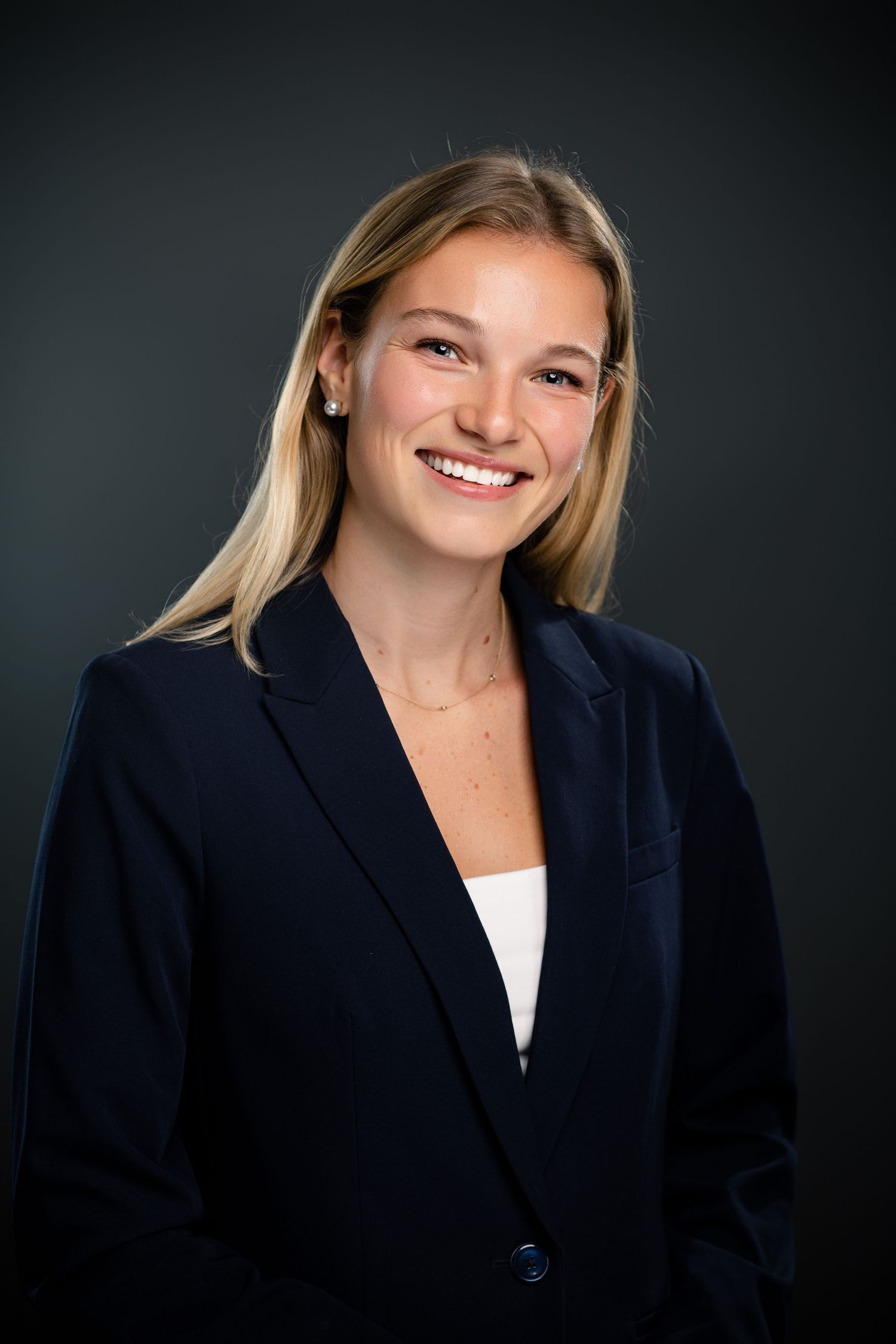 Young professional woman in navy blazer smiling confidently against dark background in corporate headshot photo.