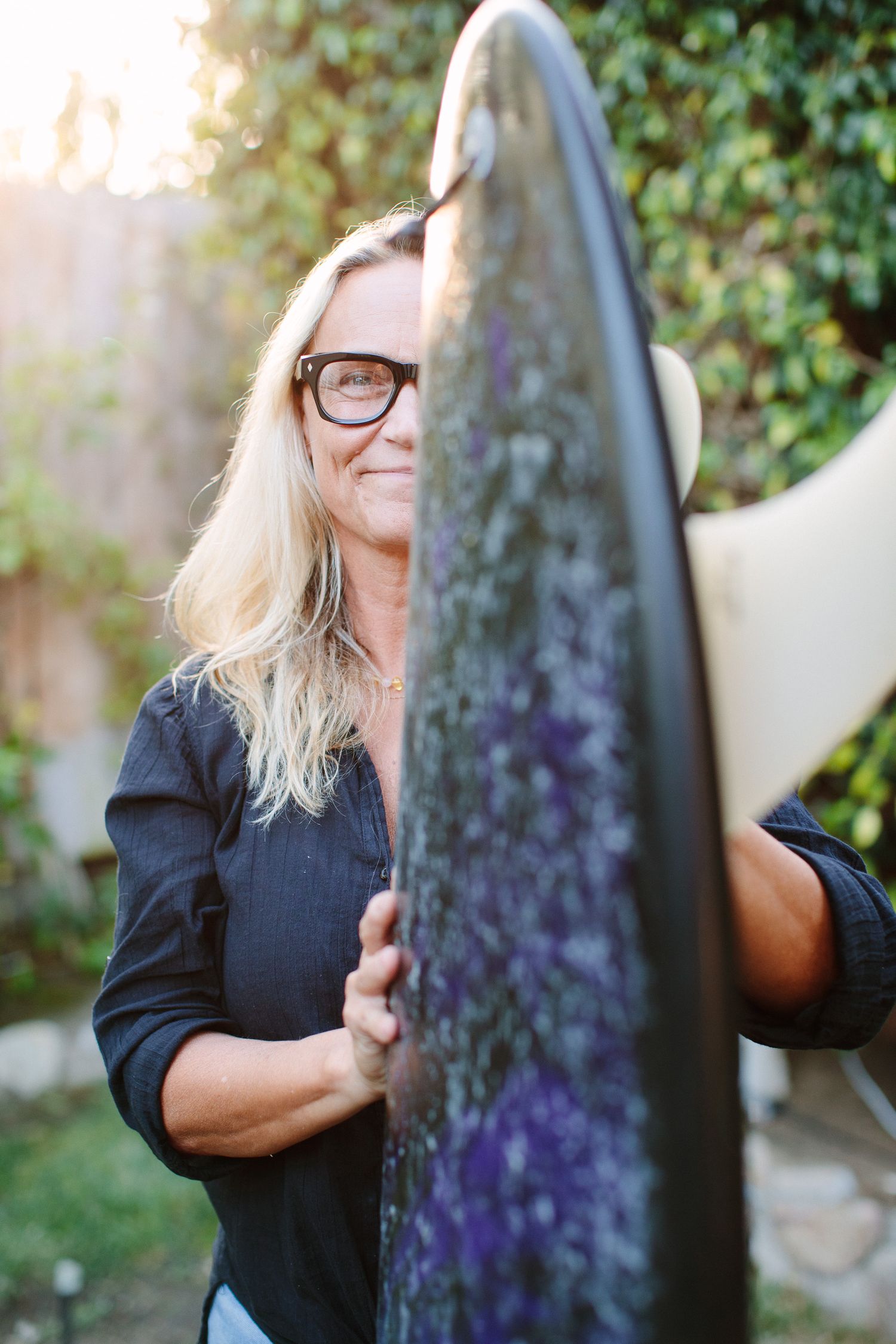 A person wearing glasses and a black shirt holds up a large dark surfboard in a sunny outdoor garden setting.