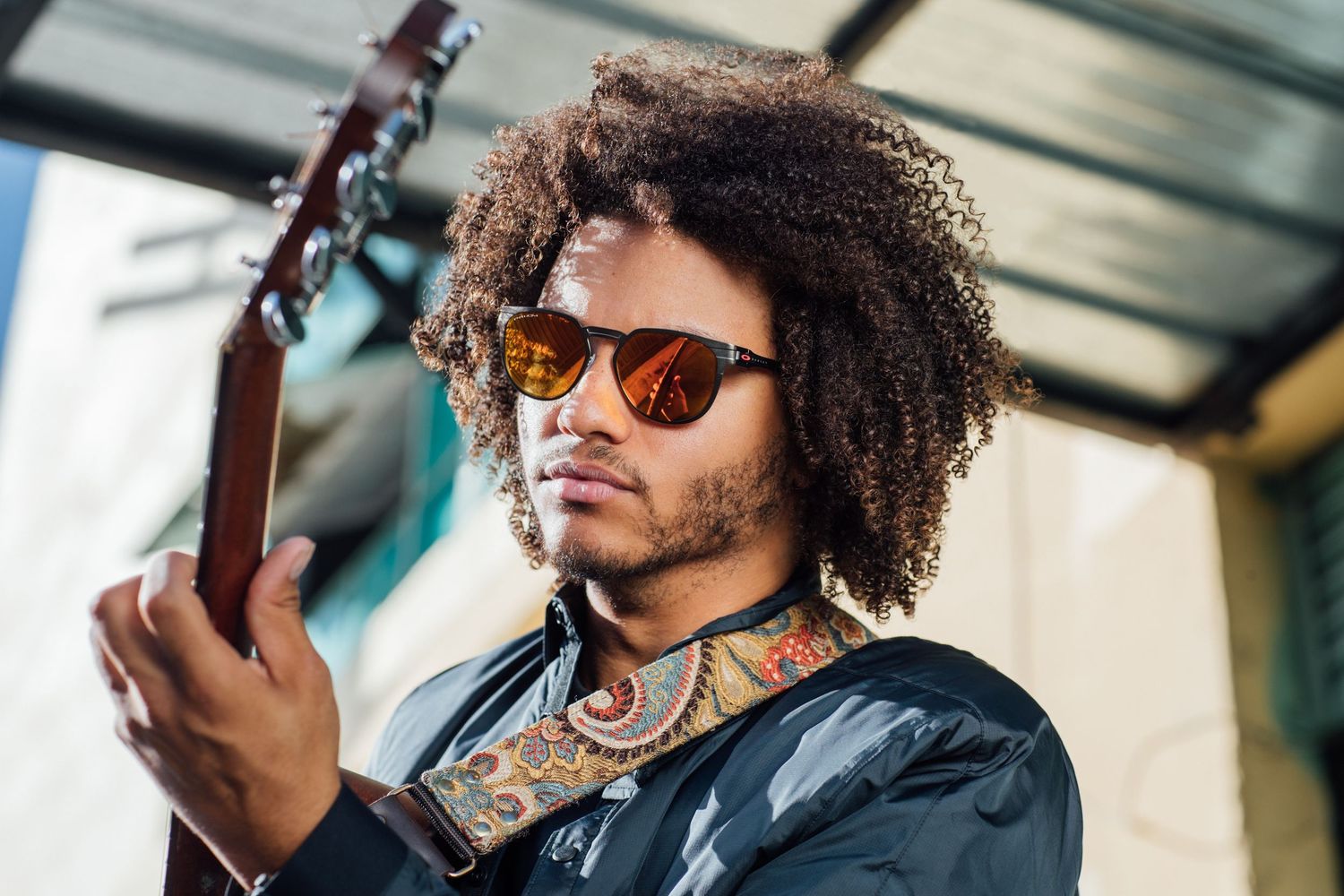 A musician with curly hair and sunglasses plays guitar while wearing a dark jacket and colorful neck tattoos.