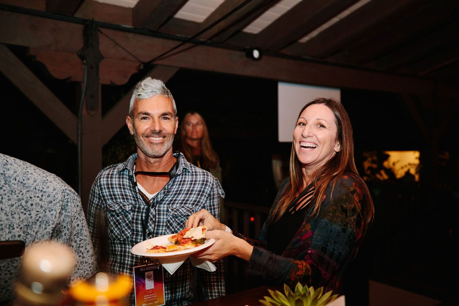 Two people share food and laughter at a casual evening gathering under wooden beams.