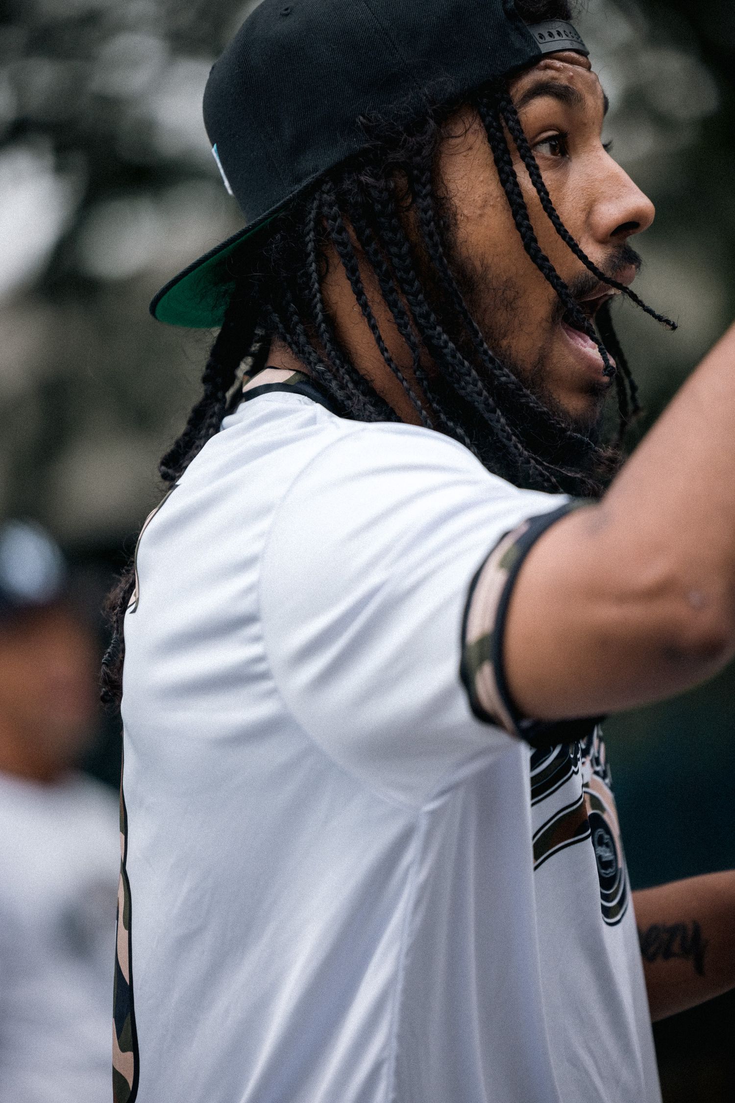 A side profile view of someone in a white t-shirt and black baseball cap with braided hair.