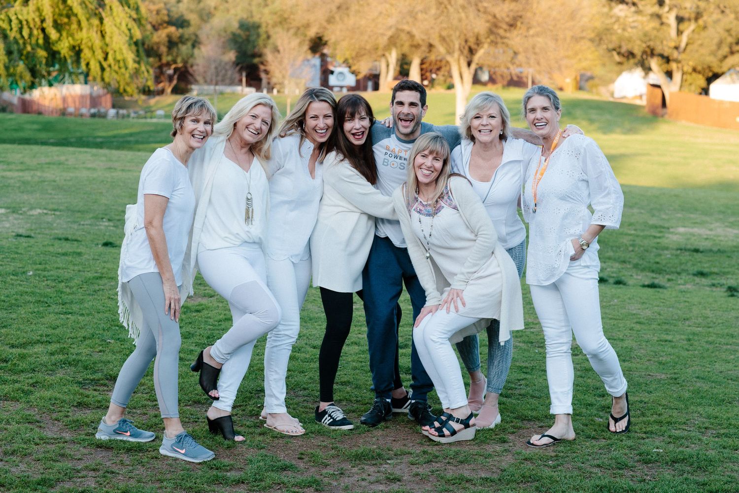 A group in all-white outfits poses playfully together on a green lawn during an outdoor photoshoot at sunset.